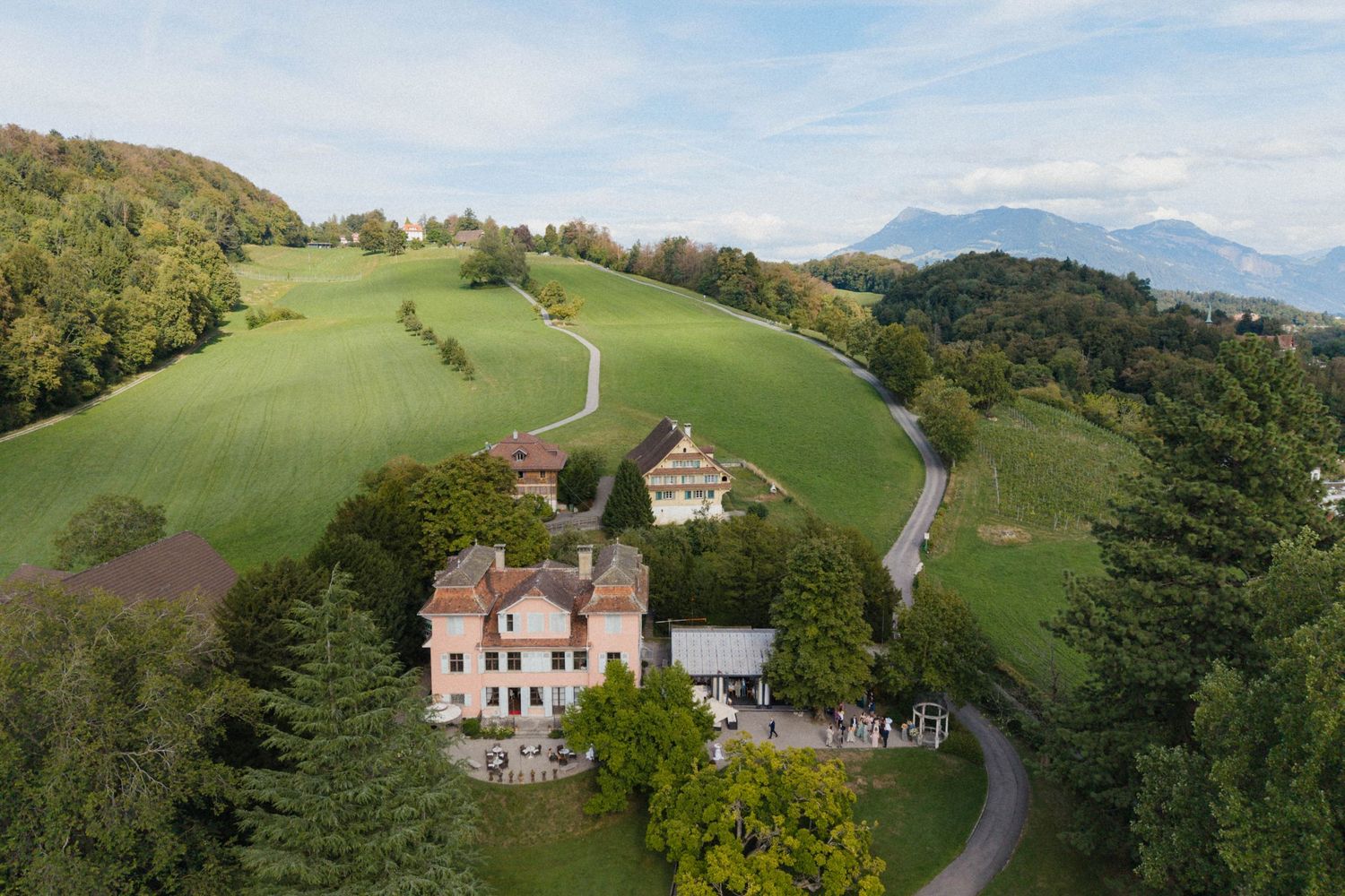 Aerial view of a historic villa surrounded by lush green hills and trees with mountain views in the background.