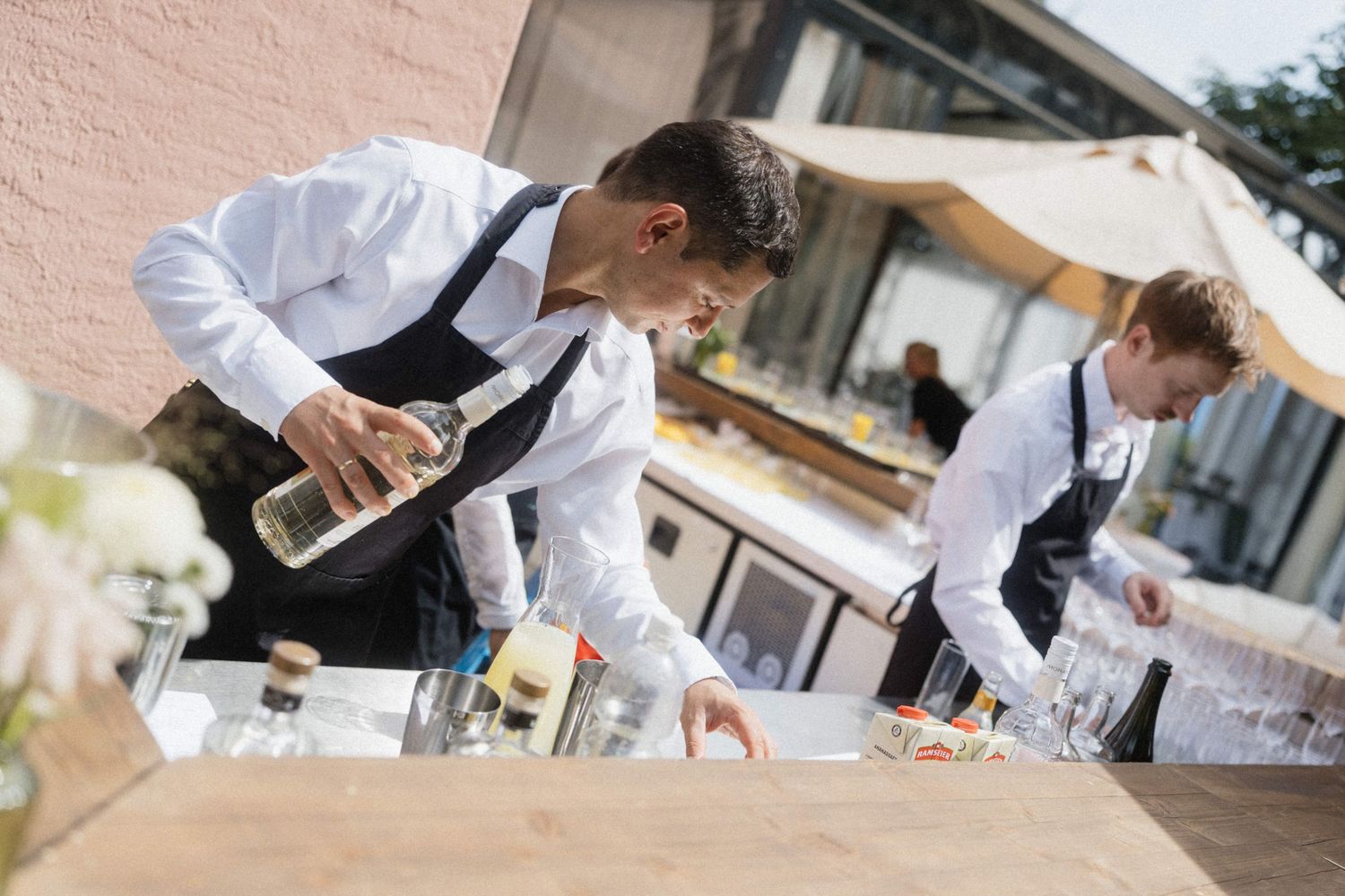 Two professional waiters in white shirts and black aprons setting up an outdoor bar event with glassware.