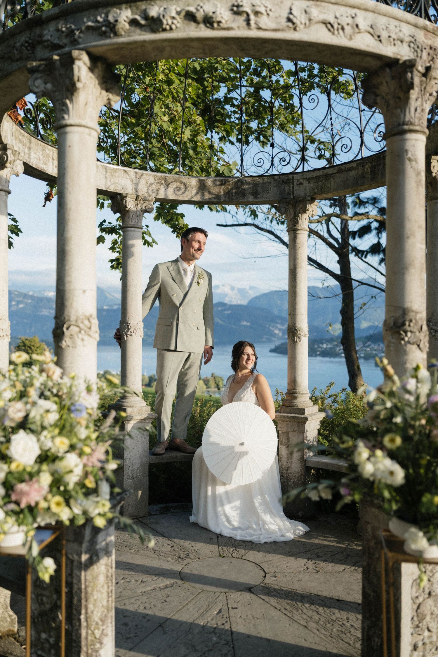Bride and groom posing inside ornate stone gazebo with alpine lake views.