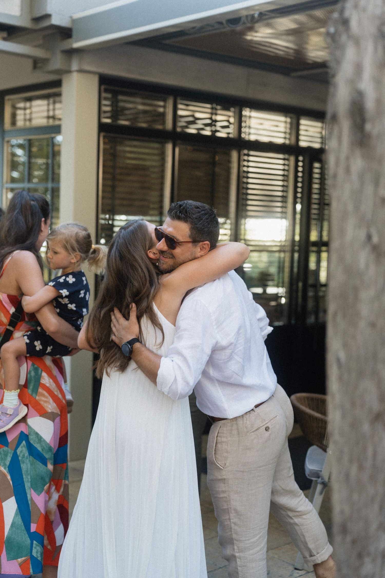 Couple embracing and kissing at an outdoor wedding reception, woman in white dress, man in white shirt and khaki pants.