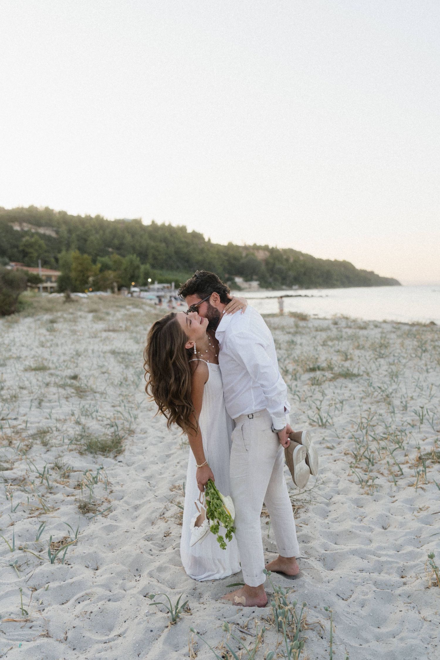 Couple kissing on white sand beach, woman in white dress holding green bouquet, man in white shirt and beige pants.
