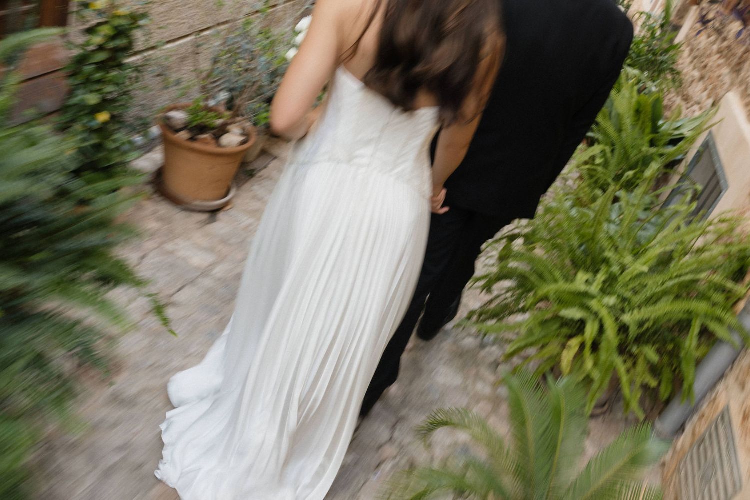 Bride in a flowing white gown walks beside a groom in a dark suit along a stone path lined with green plants and terracotta pots.