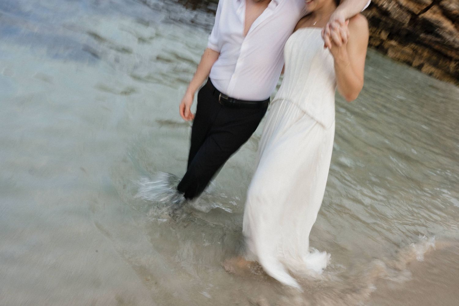 Couple in a white dress and dark trousers with white shirt walking arm in arm through shallow ocean water along a rocky beach.