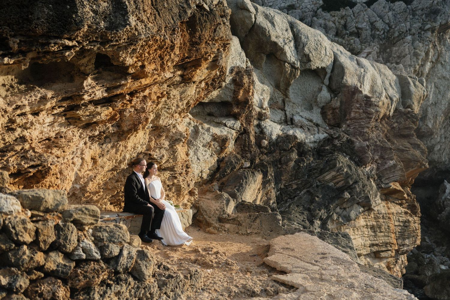 Newlyweds sit together on a sunlit rock ledge surrounded by dramatic golden cliffs during their coastal wedding photoshoot.