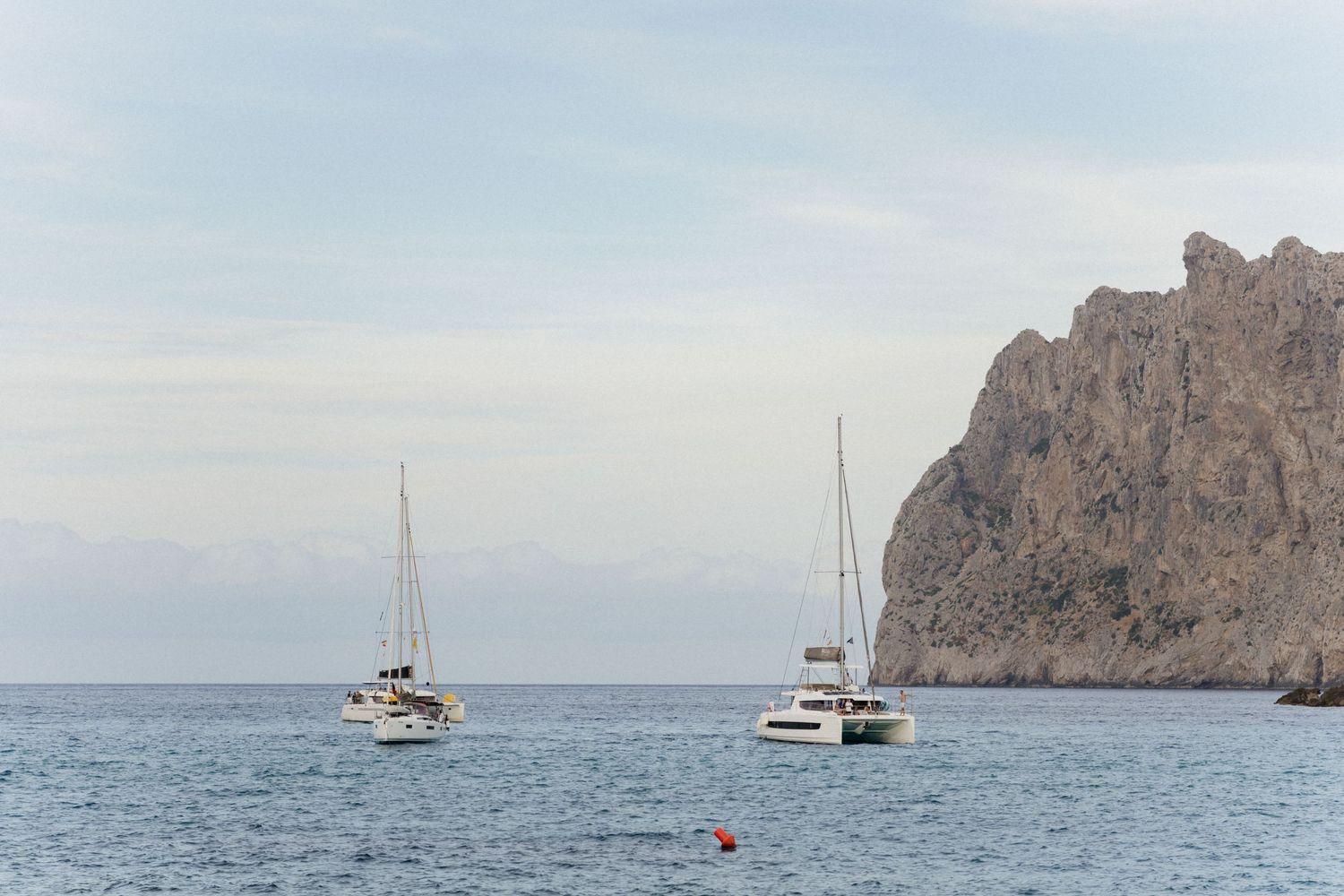 Two white sailboats anchored on calm blue sea beside a rugged rocky cliff under a soft cloudy sky.