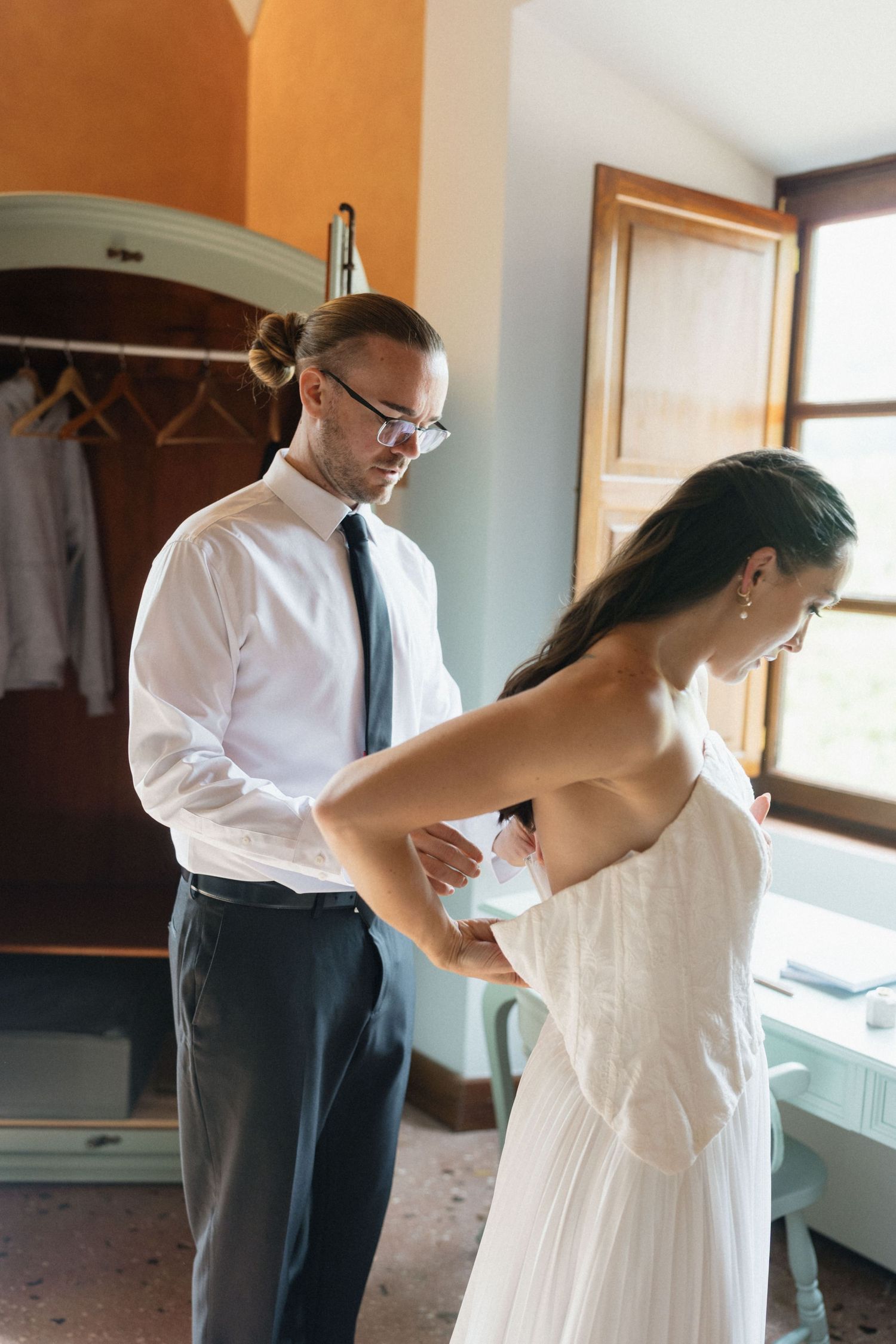 Groom in glasses and a tie helps button the back of the bride's white strapless wedding dress in a bright room.