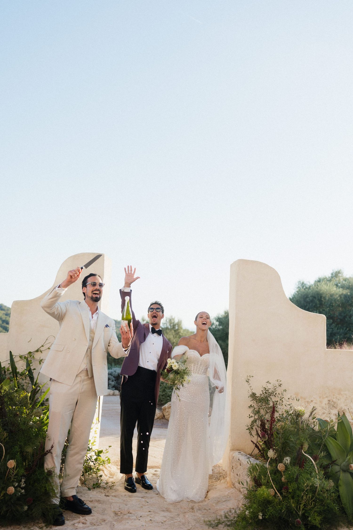 A groom in a cream suit raises a sword, a man in a burgundy blazer holds champagne, and a bride in white cheers at a sunny outdoor wedding.