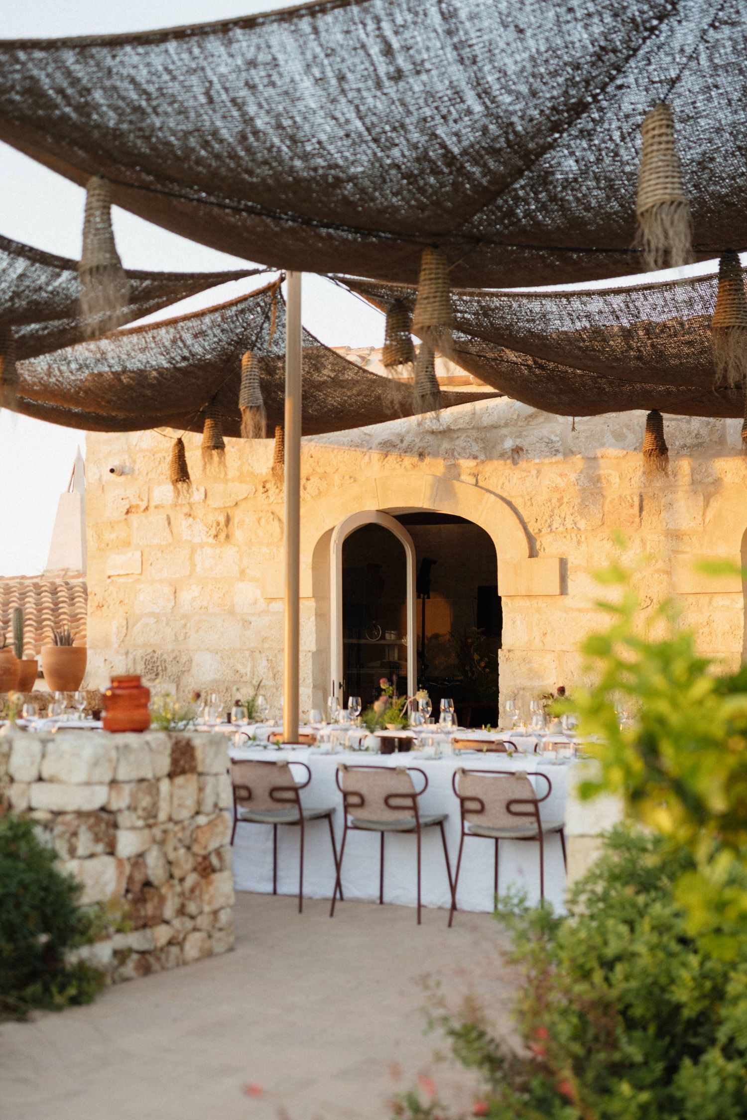 Elegant outdoor wedding dinner table beneath woven shade canopies in front of a stone arched building.