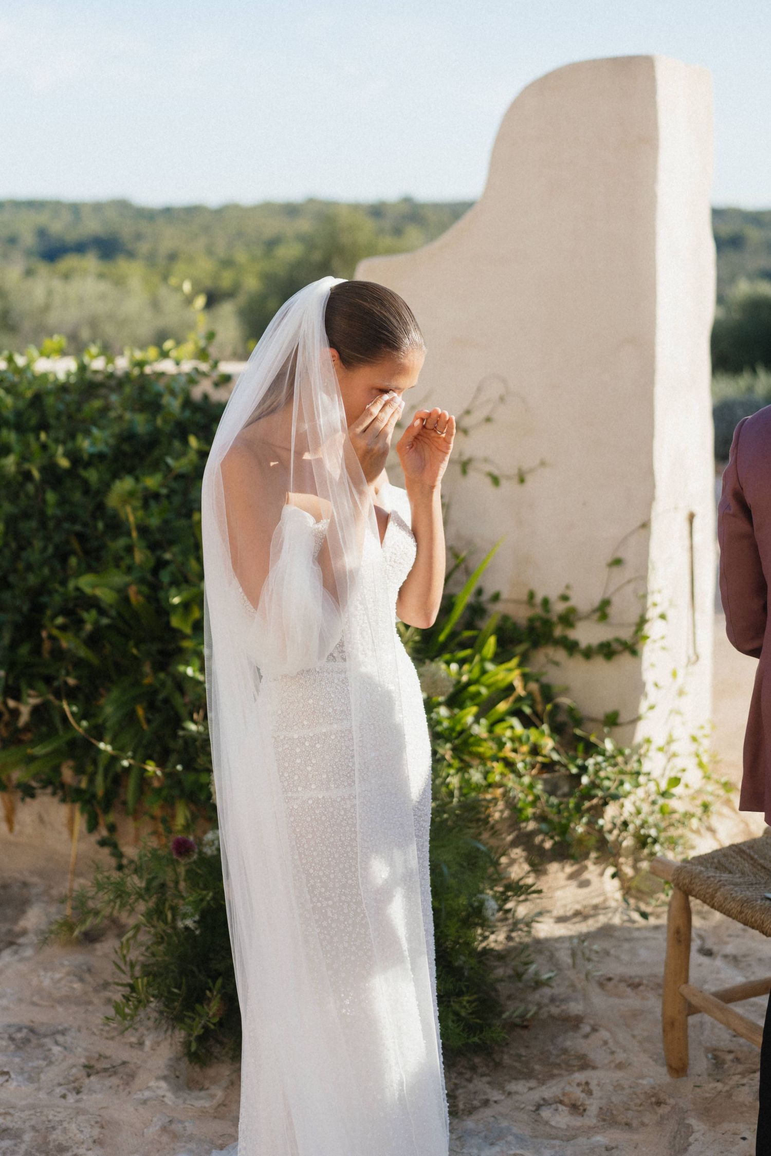 Bride in a long veil and white gown wipes tears from her face during an emotional outdoor wedding ceremony.