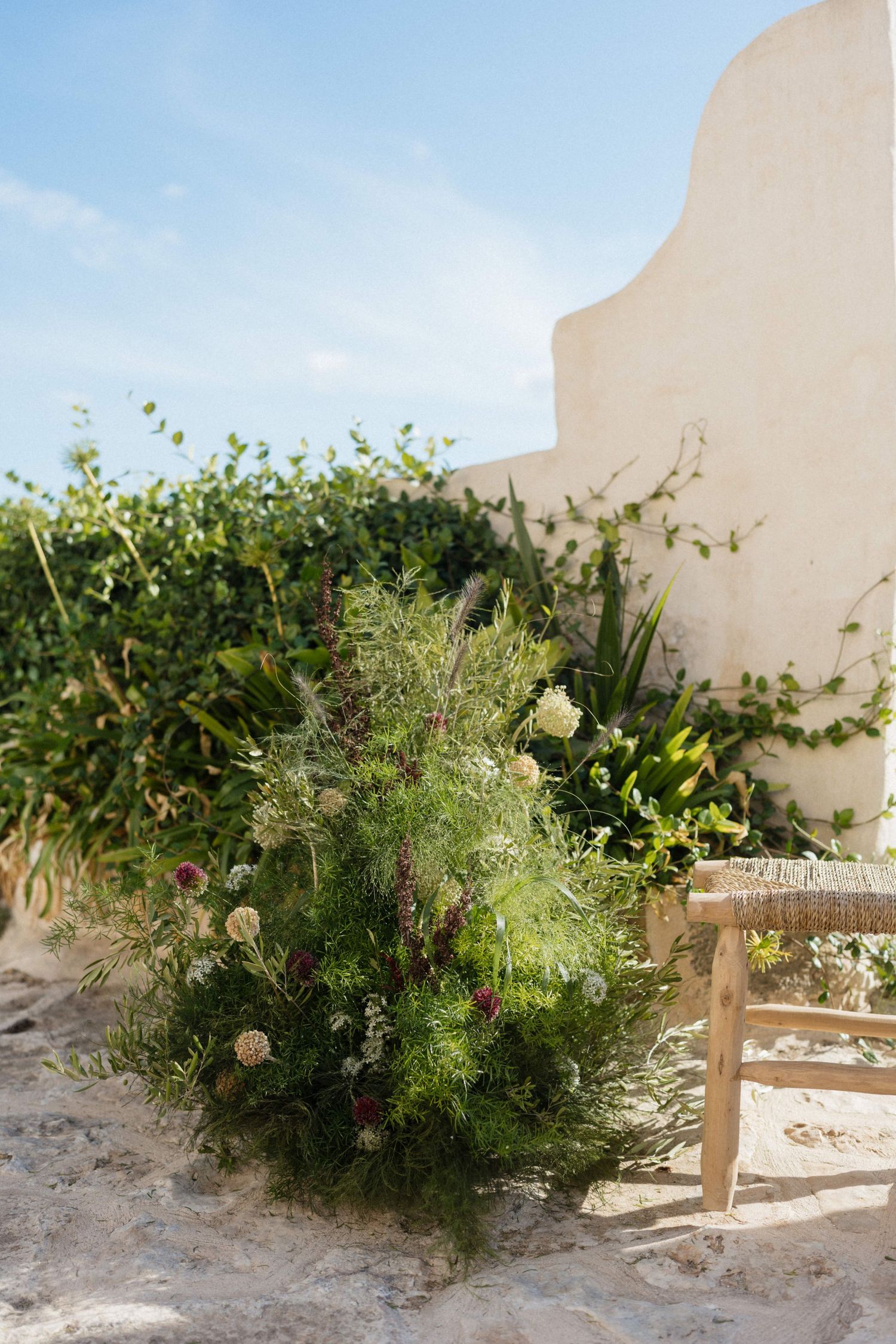 Wild floral arrangement with greenery and blooms sits beside a rustic wooden chair near a whitewashed wall.