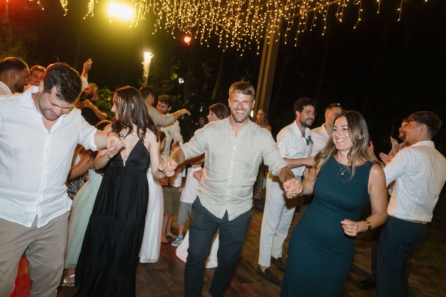 Guests dancing joyfully at an outdoor wedding reception under warm string lights at night.