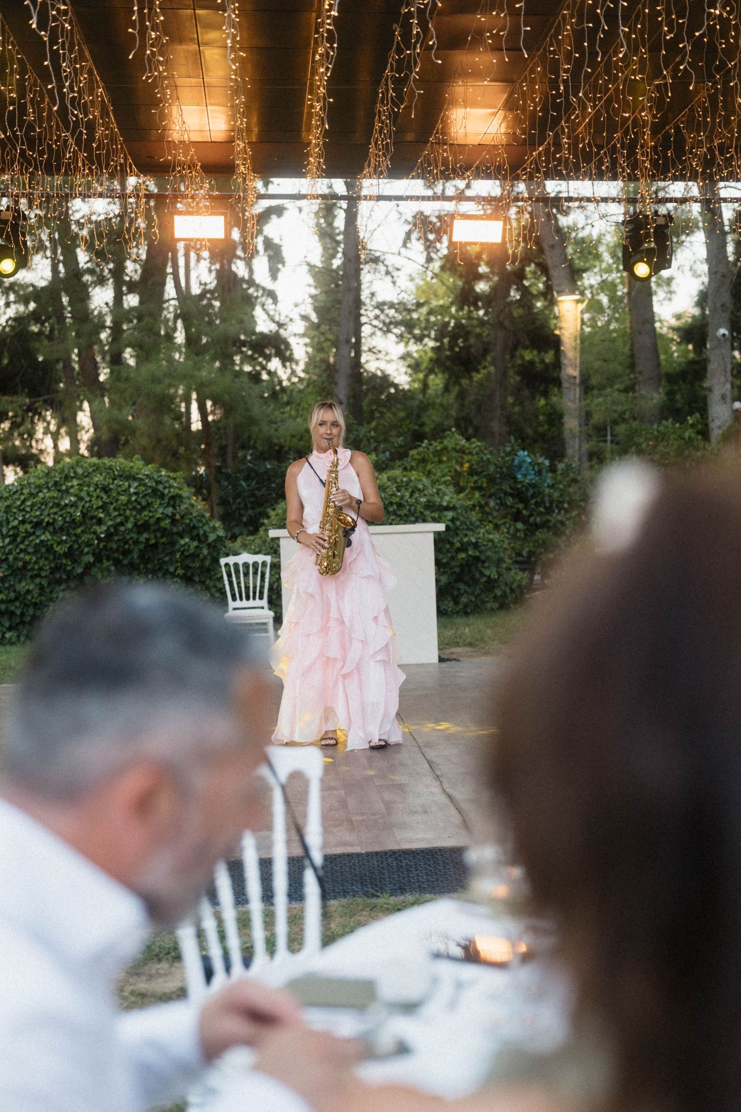 A woman in a pink gown plays saxophone at an elegant outdoor wedding reception under string lights.