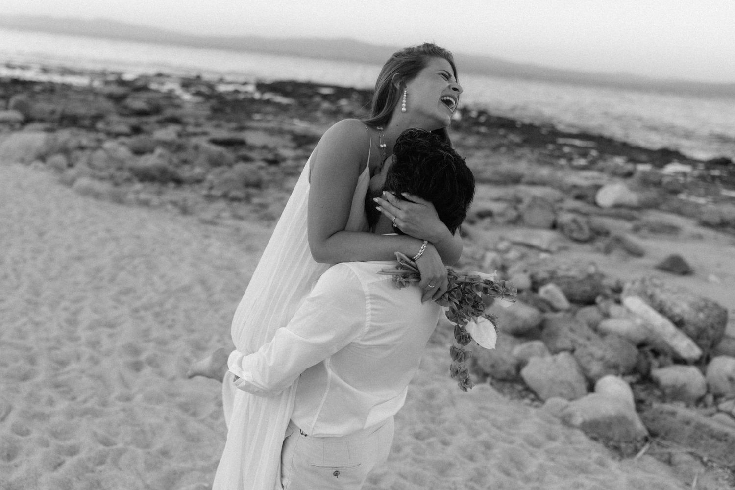 Black and white photo of bride laughing while embracing groom on sandy beach with rocks behind them.