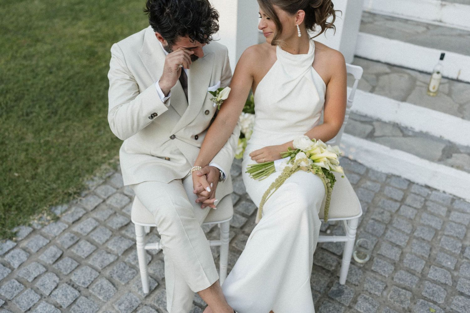 Bride and groom sitting together on white chairs, smiling and holding hands, dressed in elegant white wedding attire with a bridal bouquet.