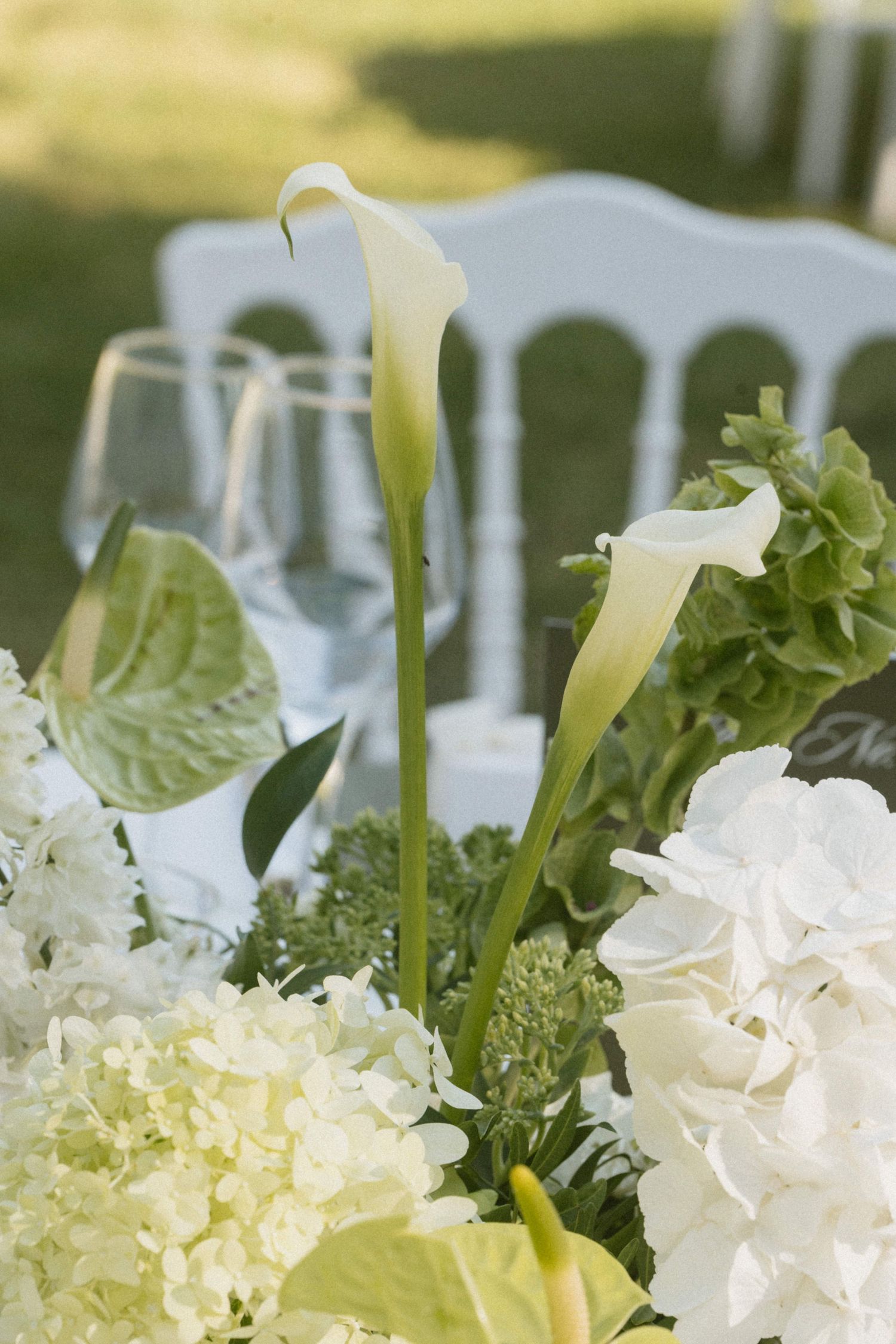 White calla lilies and hydrangeas centerpiece with wine glasses on an outdoor wedding reception table.