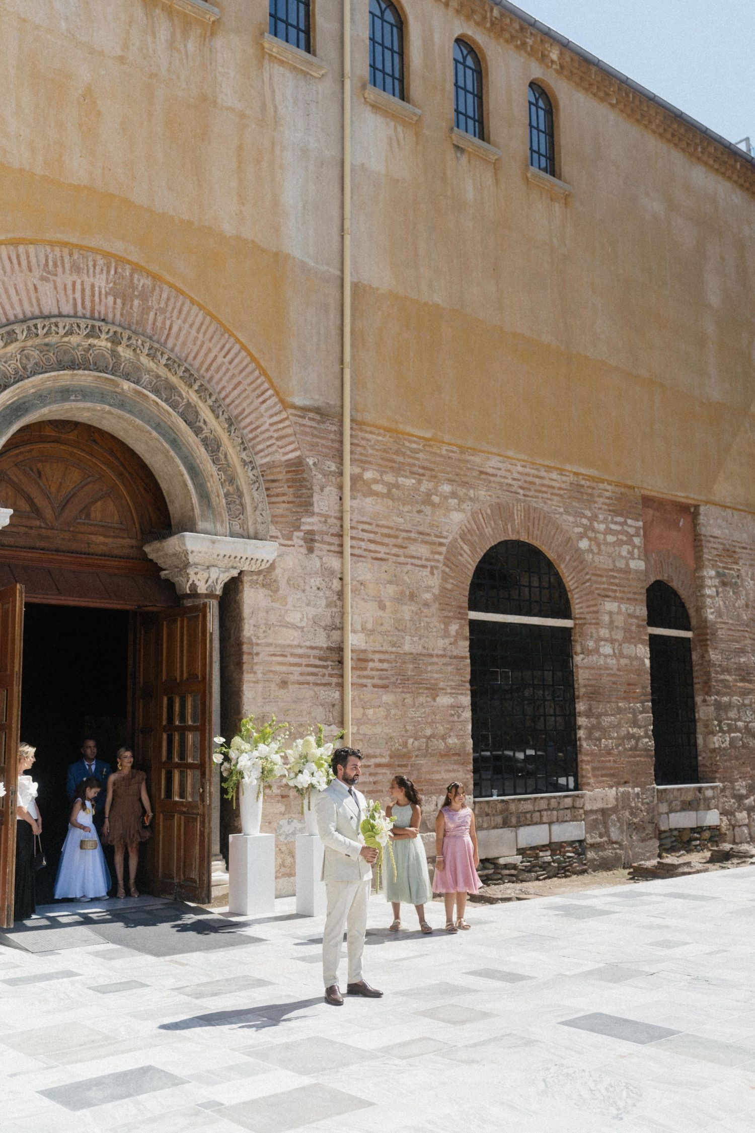 Groom in ivory suit holding bouquet outside ornate Byzantine stone church on sunny wedding day.