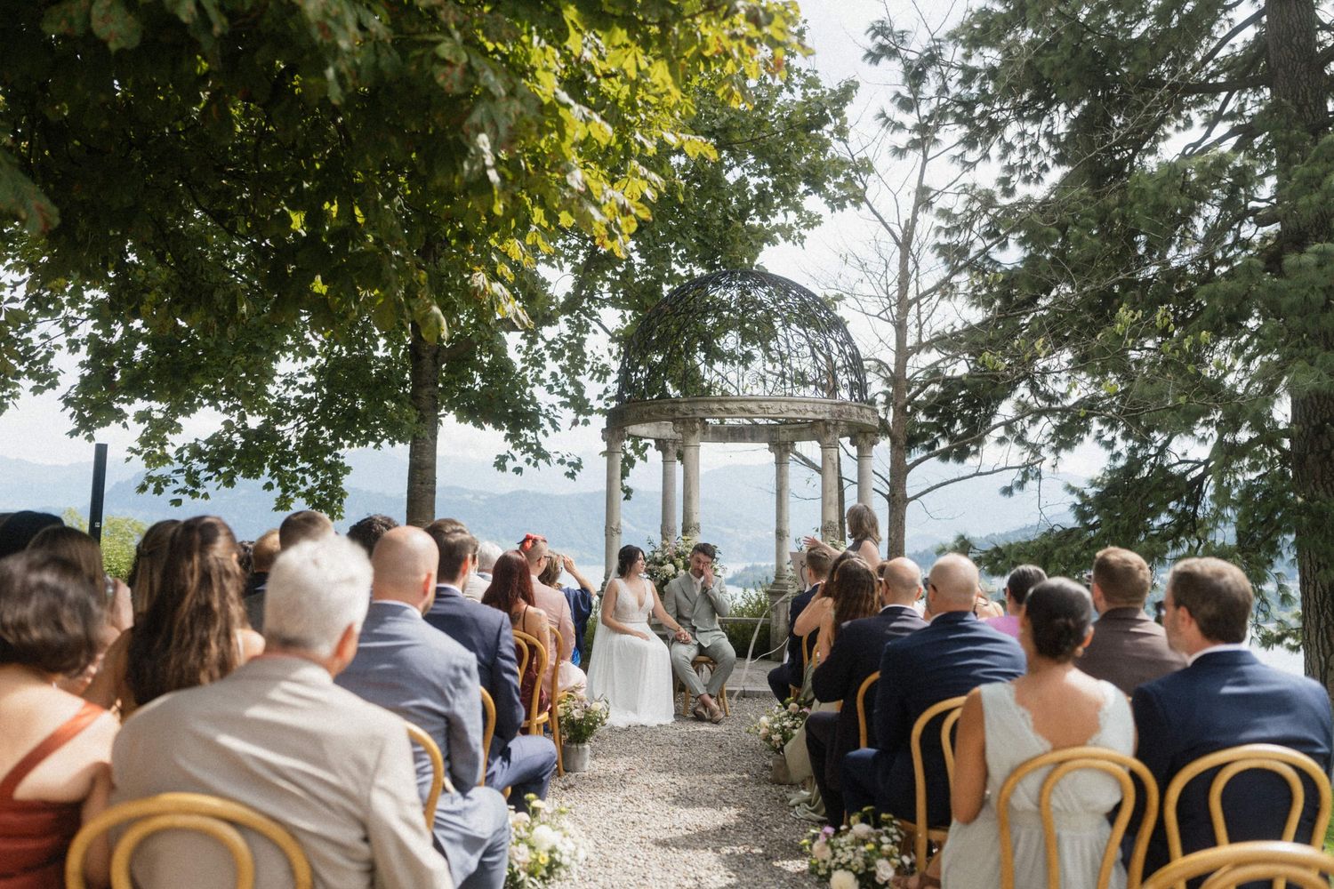 Outdoor wedding ceremony with guests seated facing couple at a gazebo overlooking a scenic lake and mountains.