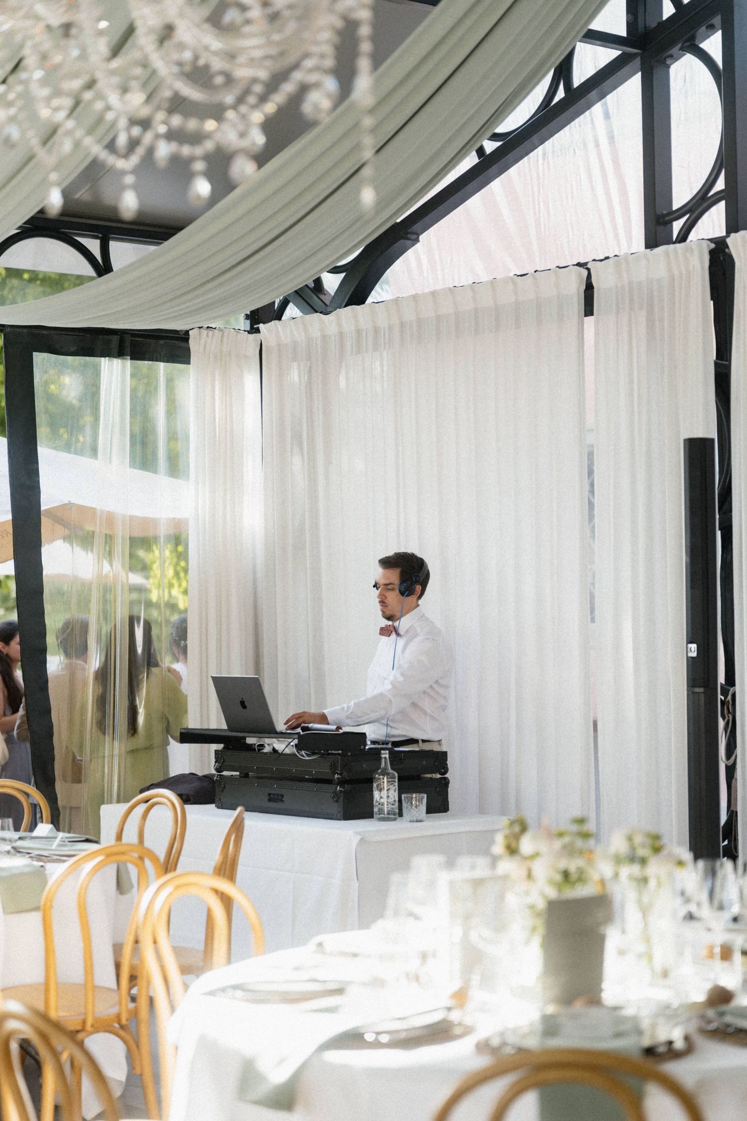 DJ in white shirt sets up equipment in an elegant draped reception tent with chandelier and wooden chairs.