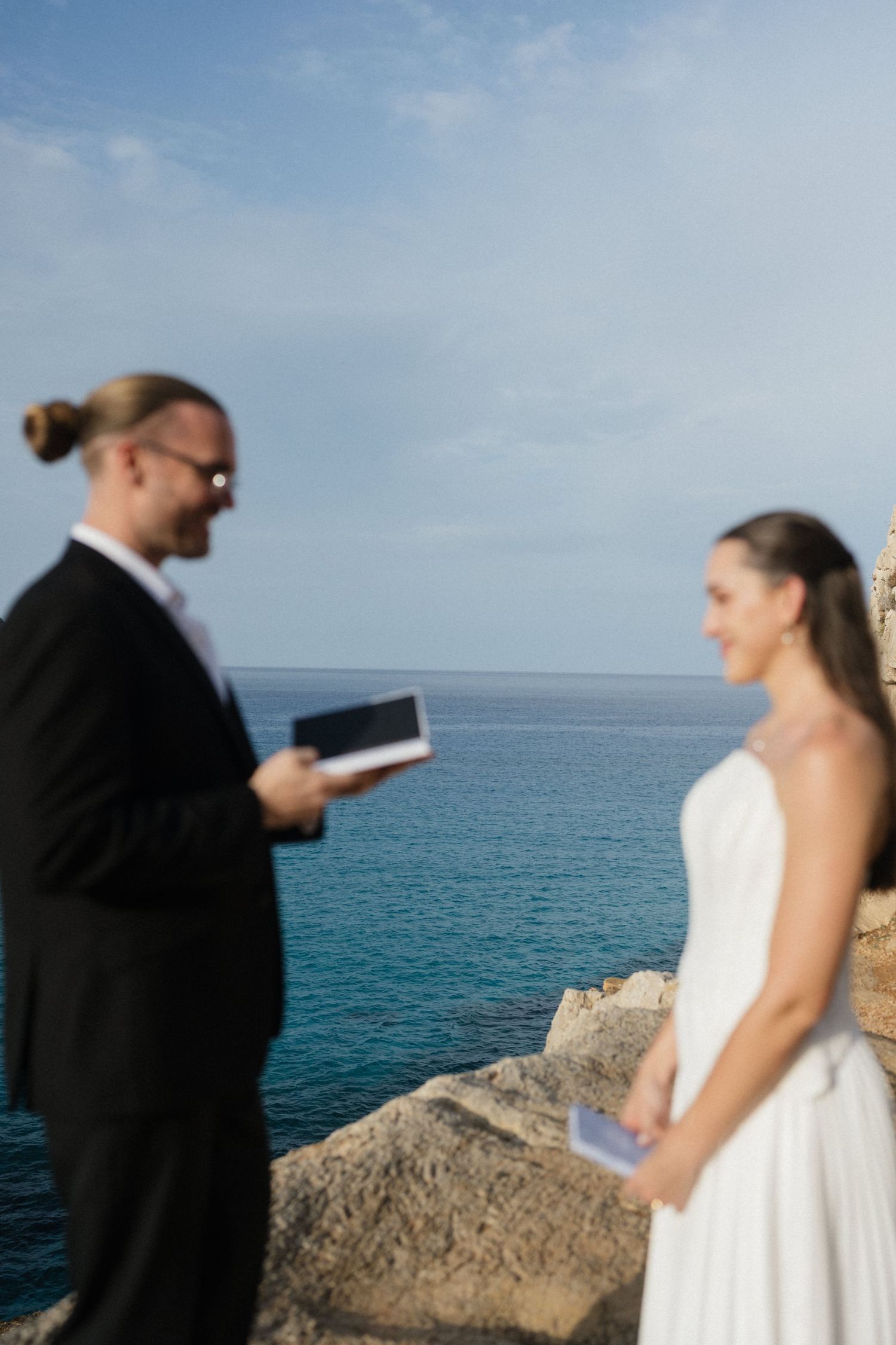 A groom with a bun and glasses reads from a book while a bride in a white gown listens during a seaside cliff wedding ceremony.