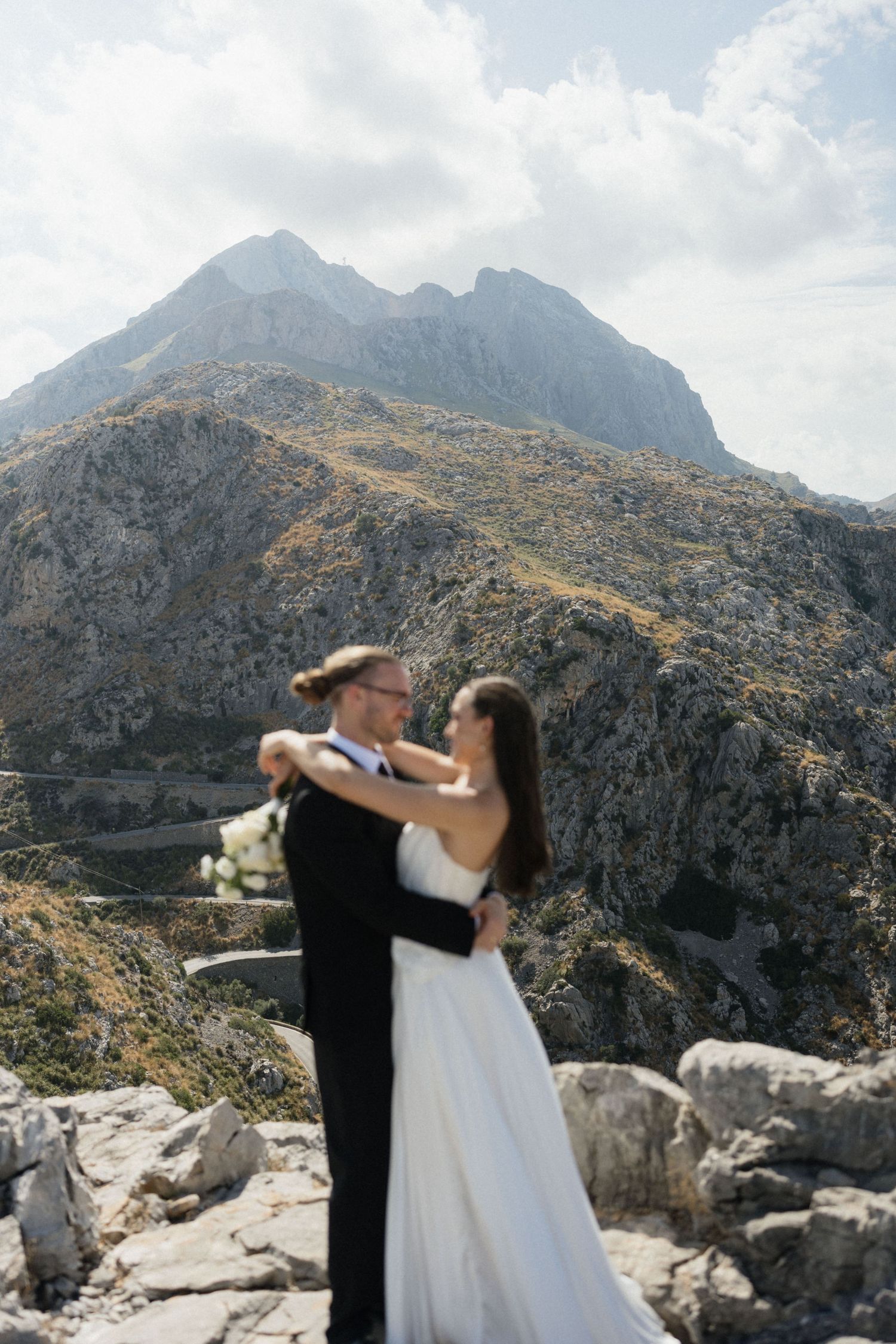 Bride in a white gown and groom in a dark suit embrace on a rocky mountain overlook during a scenic destination wedding photoshoot.