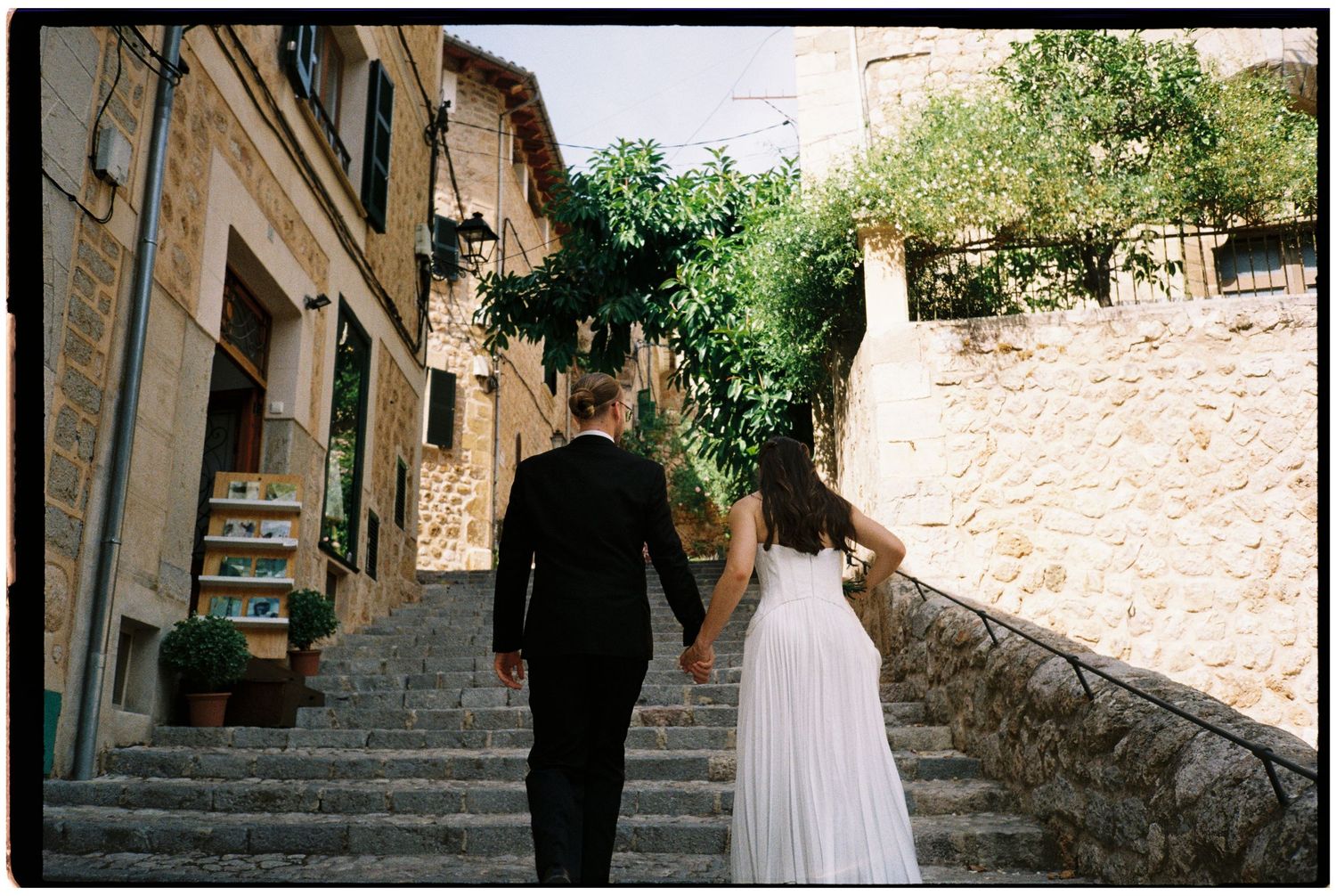Bride in a white gown and groom in a dark suit hold hands while walking up a historic stone staircase in a charming Mediterranean village.