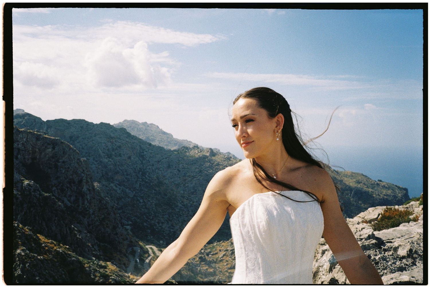 Woman in a white strapless dress stands on a rocky mountainside with her hair blowing in the wind, overlooking the sea under a blue sky.