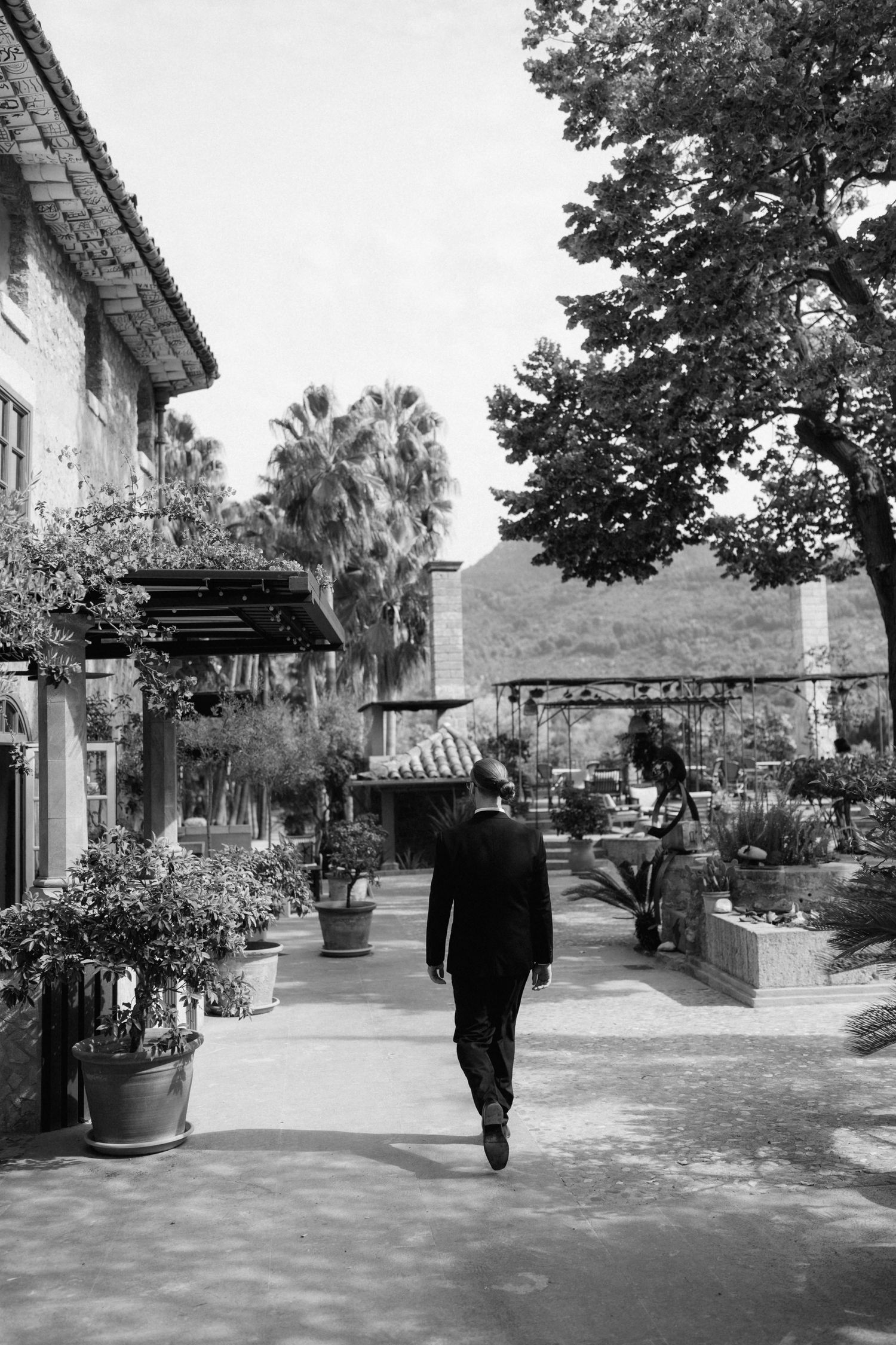 Black and white photo of a groom in a dark suit walking away along a sunlit courtyard with potted plants, palm trees and mountains behind.