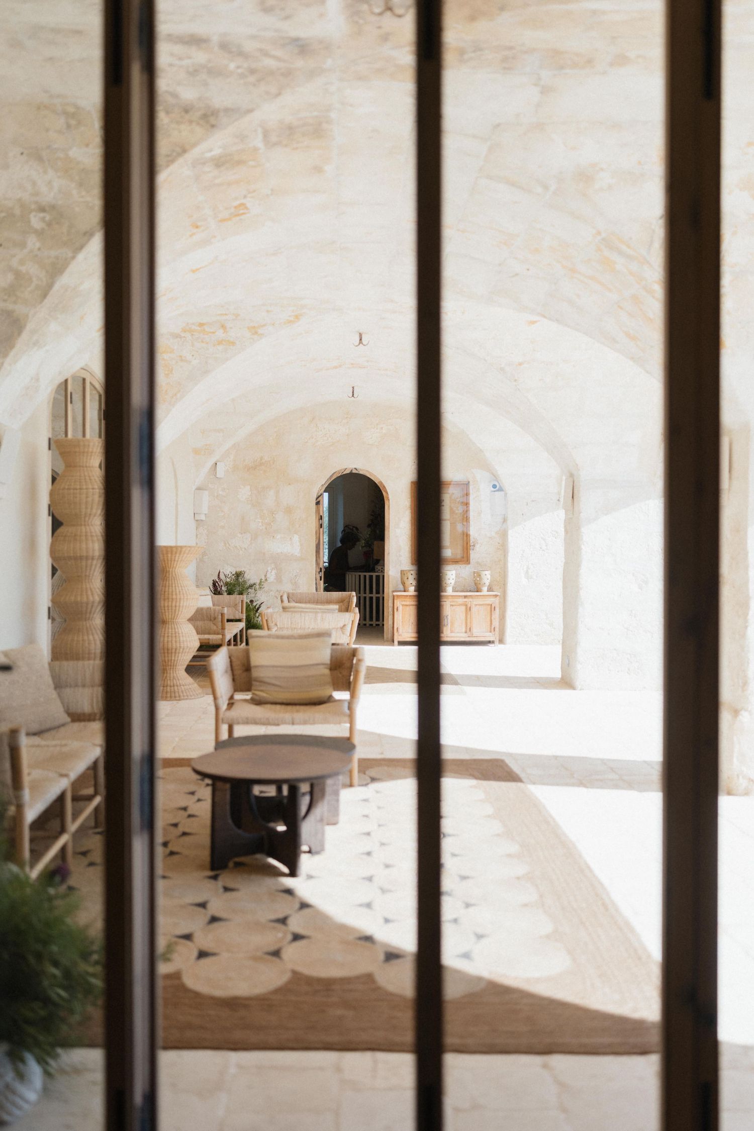 Sunlit stone-vaulted lounge with wooden chairs, a round table and a patterned rug, seen through slim dark window frames.