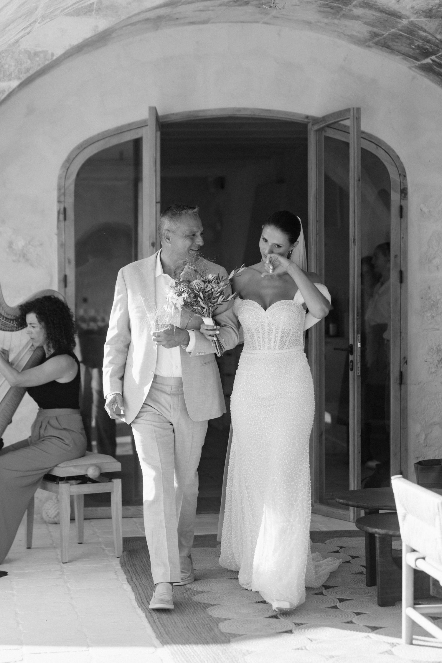 Black and white photo of a groom in a linen suit walking a tearful bride in a fitted lace gown through an arched doorway.