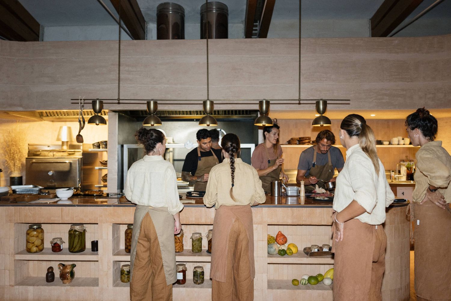 A team of chefs in aprons works together in a bright open kitchen with shelves of jars and fresh ingredients.