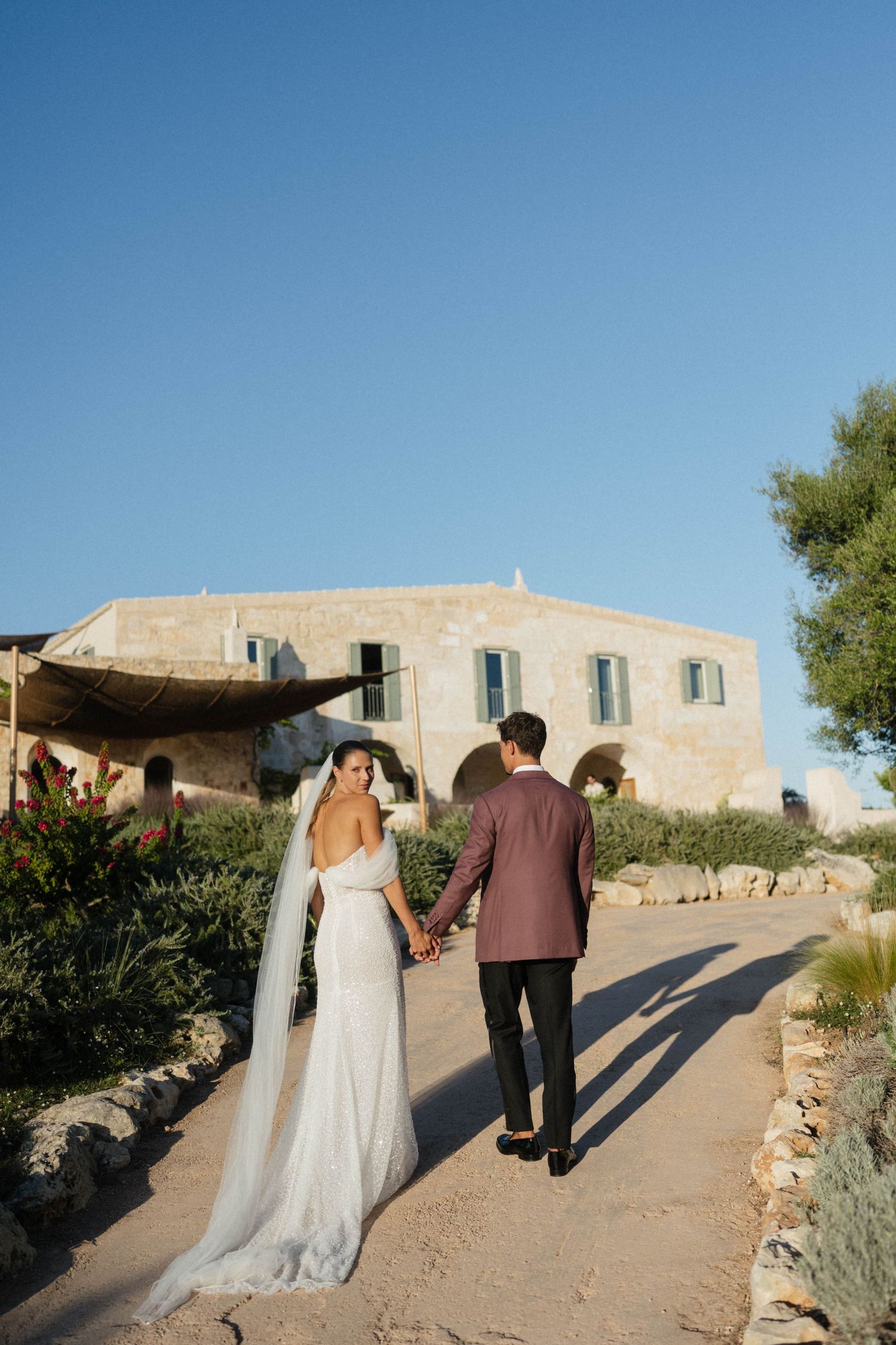 A bride in a flowing white gown and long veil walks hand in hand with her groom toward a stone villa at sunset.