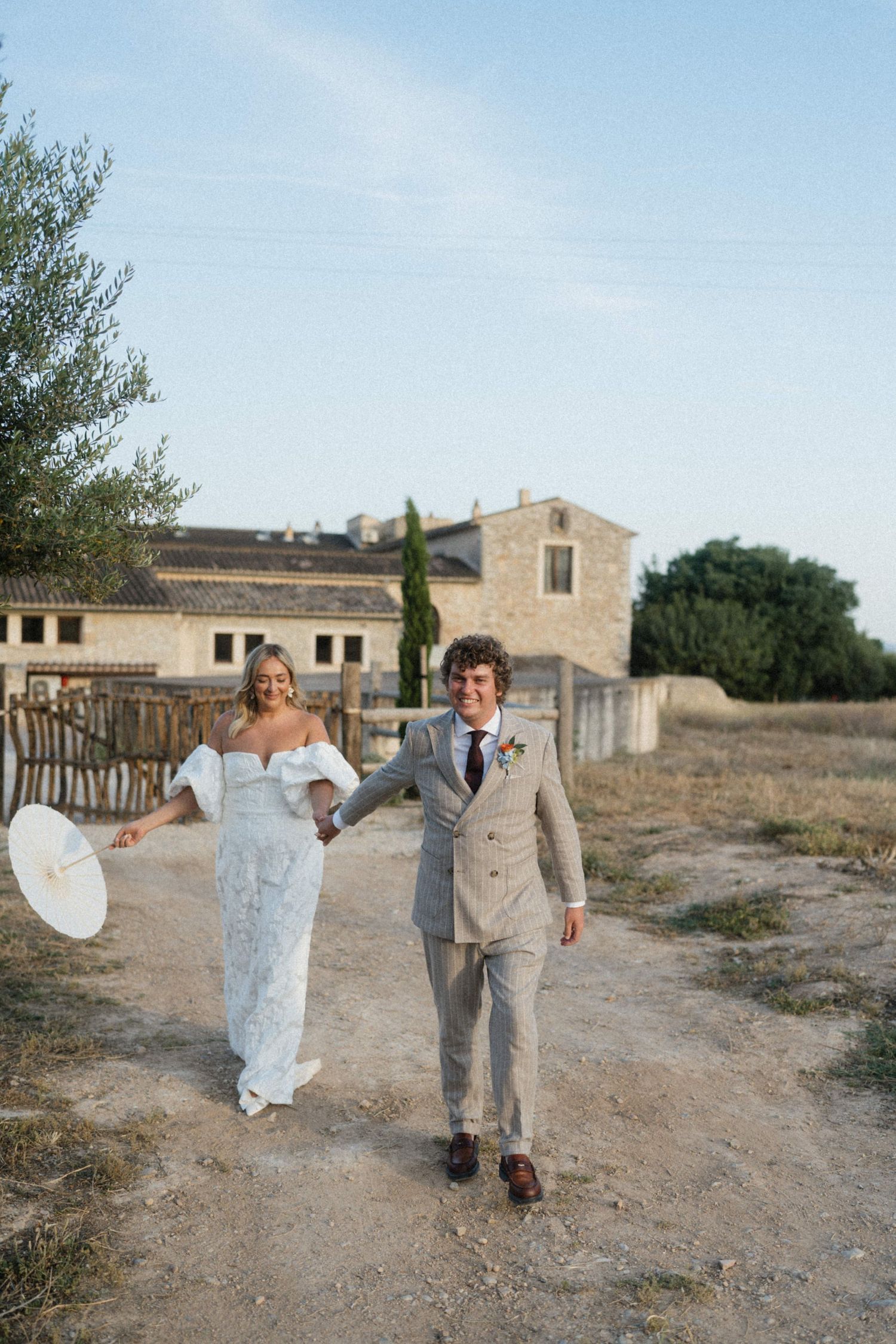Bride and groom walking hand in hand outside rustic stone farmhouse on sunny wedding day.