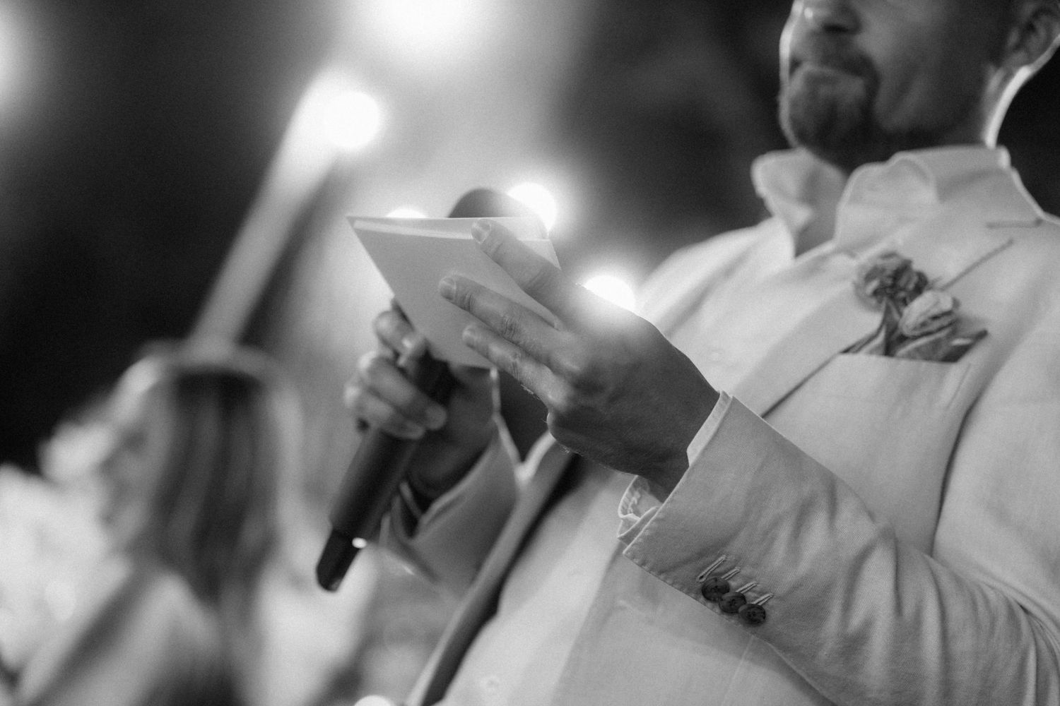 Man in white suit holding microphone and card speaks at elegant evening wedding ceremony.