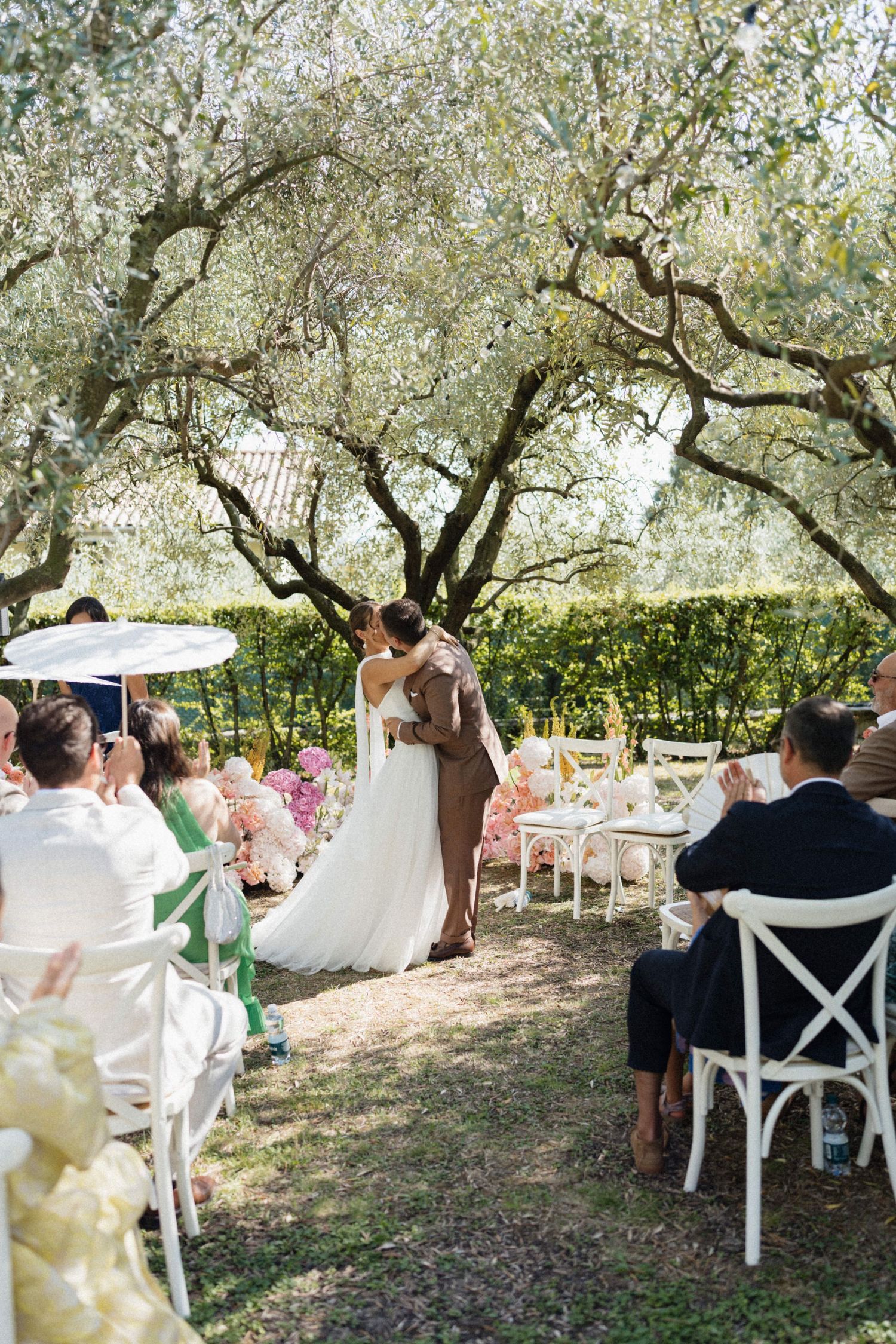 Bride and groom share first kiss under olive trees at a sunny outdoor wedding ceremony.