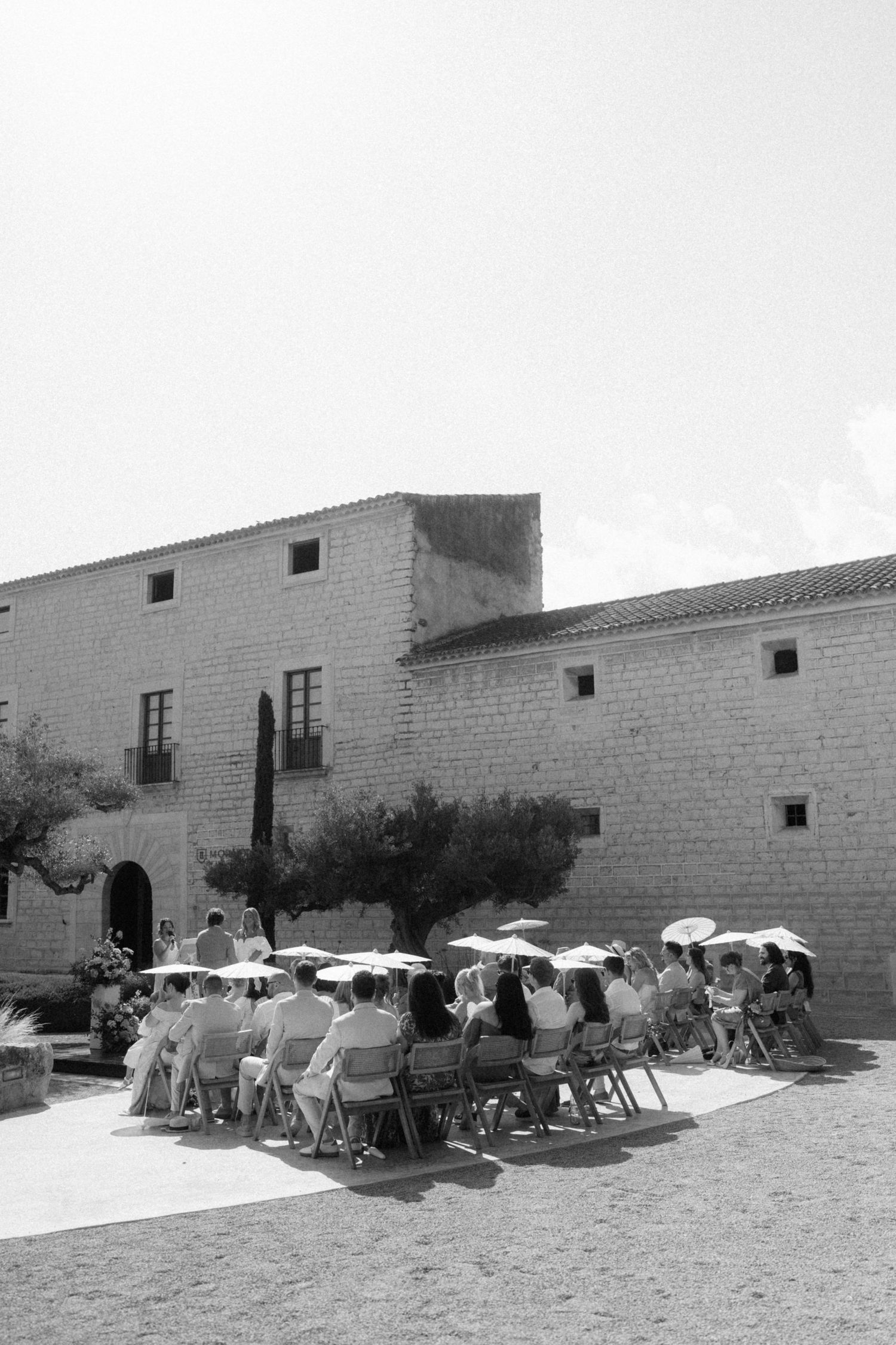 Guests seated outdoors at sunny wedding ceremony beside rustic stone building, black and white photo.