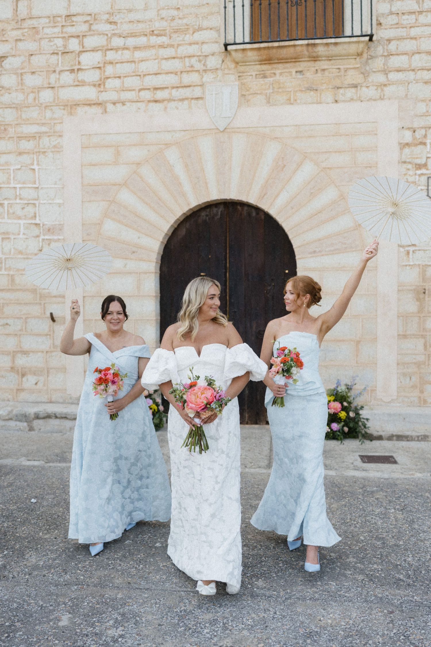 Bride and two bridesmaids in blue gowns holding paper fans outside a stone church.