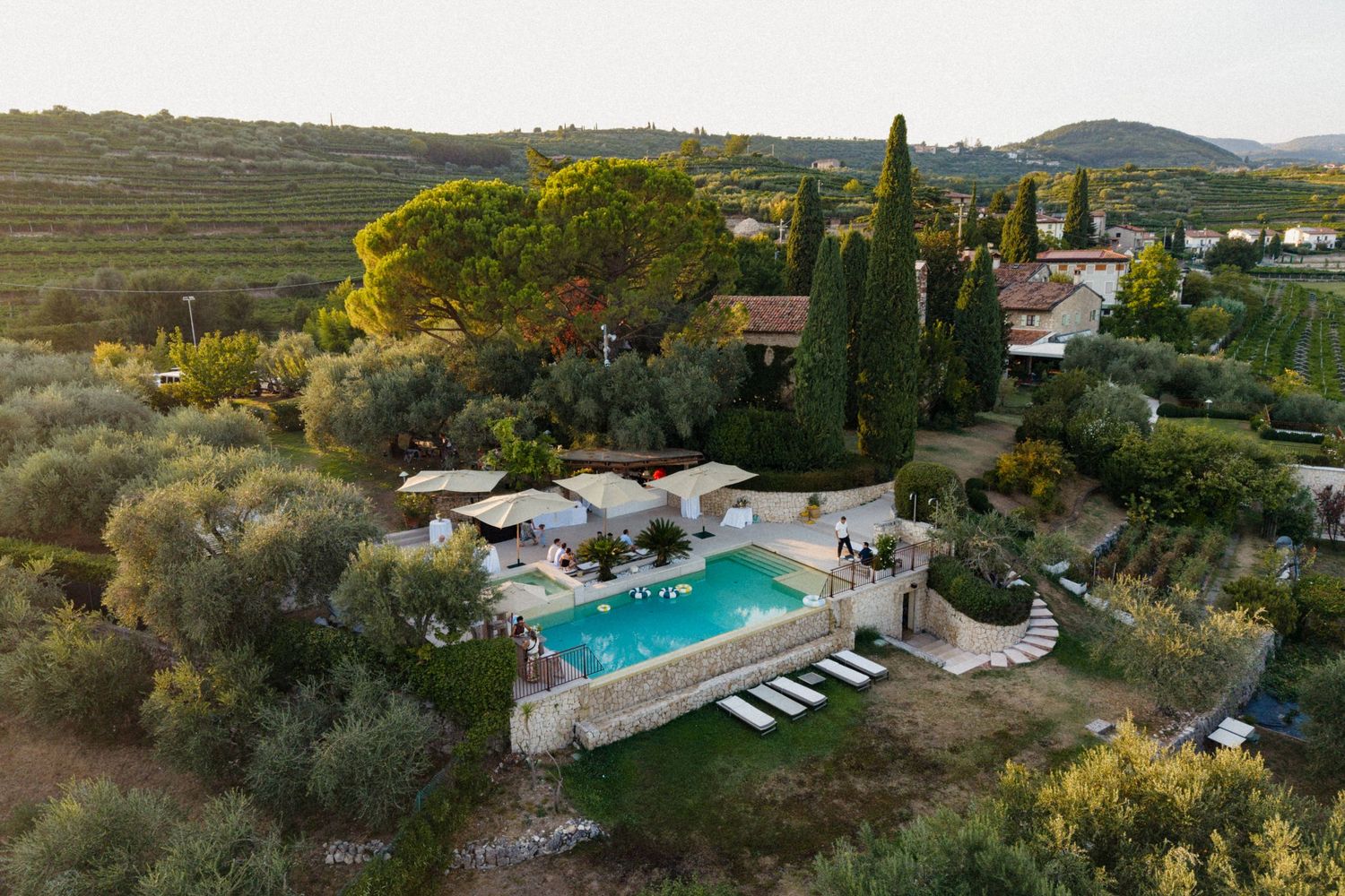 Aerial view of a Tuscan villa with a turquoise pool, surrounded by olive trees, cypresses, and rolling hills at dusk.