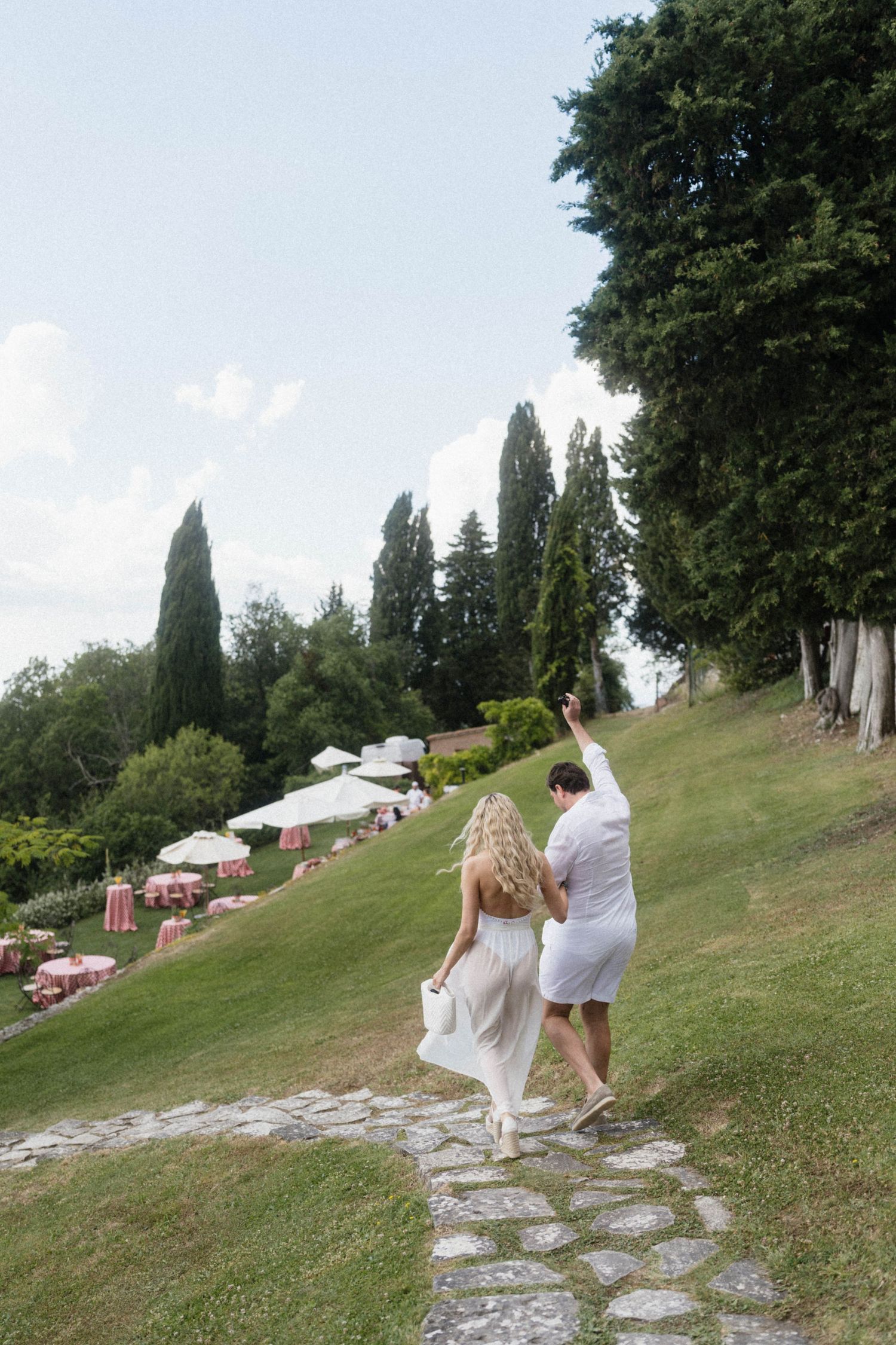 Bride and groom in white walking up a stone path through lush green gardens at a wedding.
