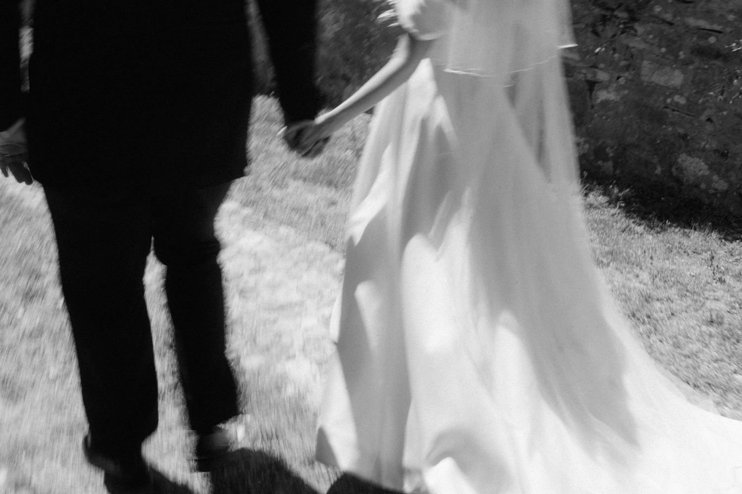 Artistic black and white photo of bride in flowing white gown holding groom's hand outdoors.