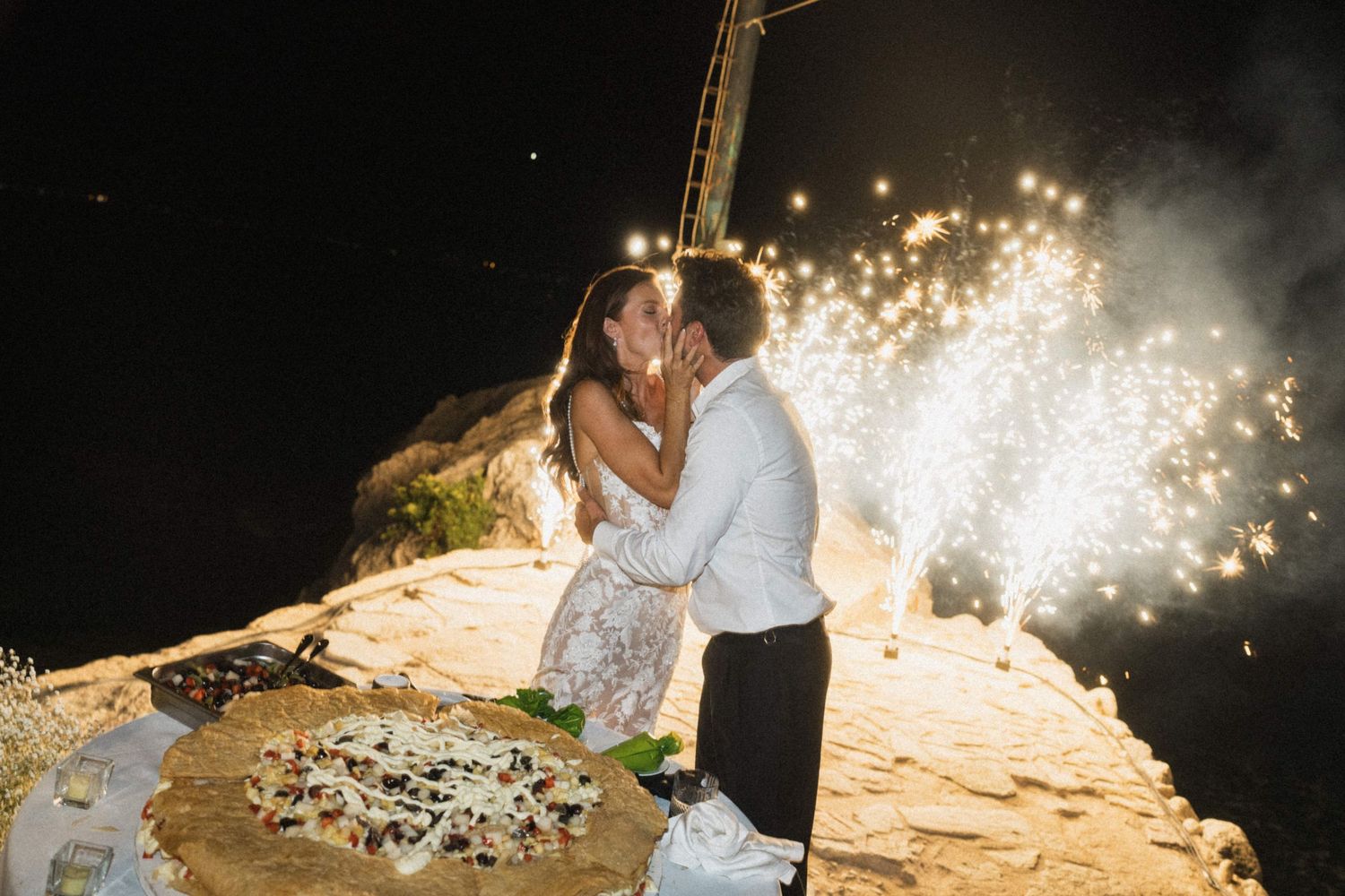 Bride and groom share romantic kiss beside wedding pizza cake with sparkling fireworks at night.