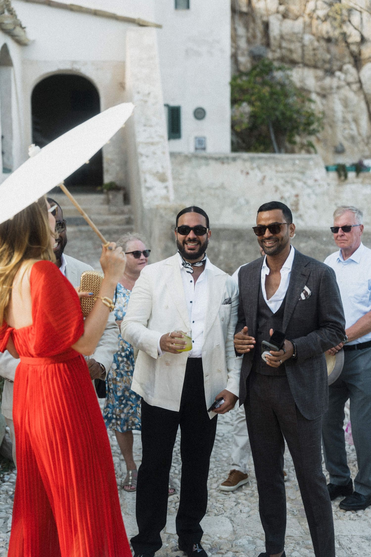 Wedding guests socializing outdoors, two stylish men in sunglasses chatting with woman in red dress.