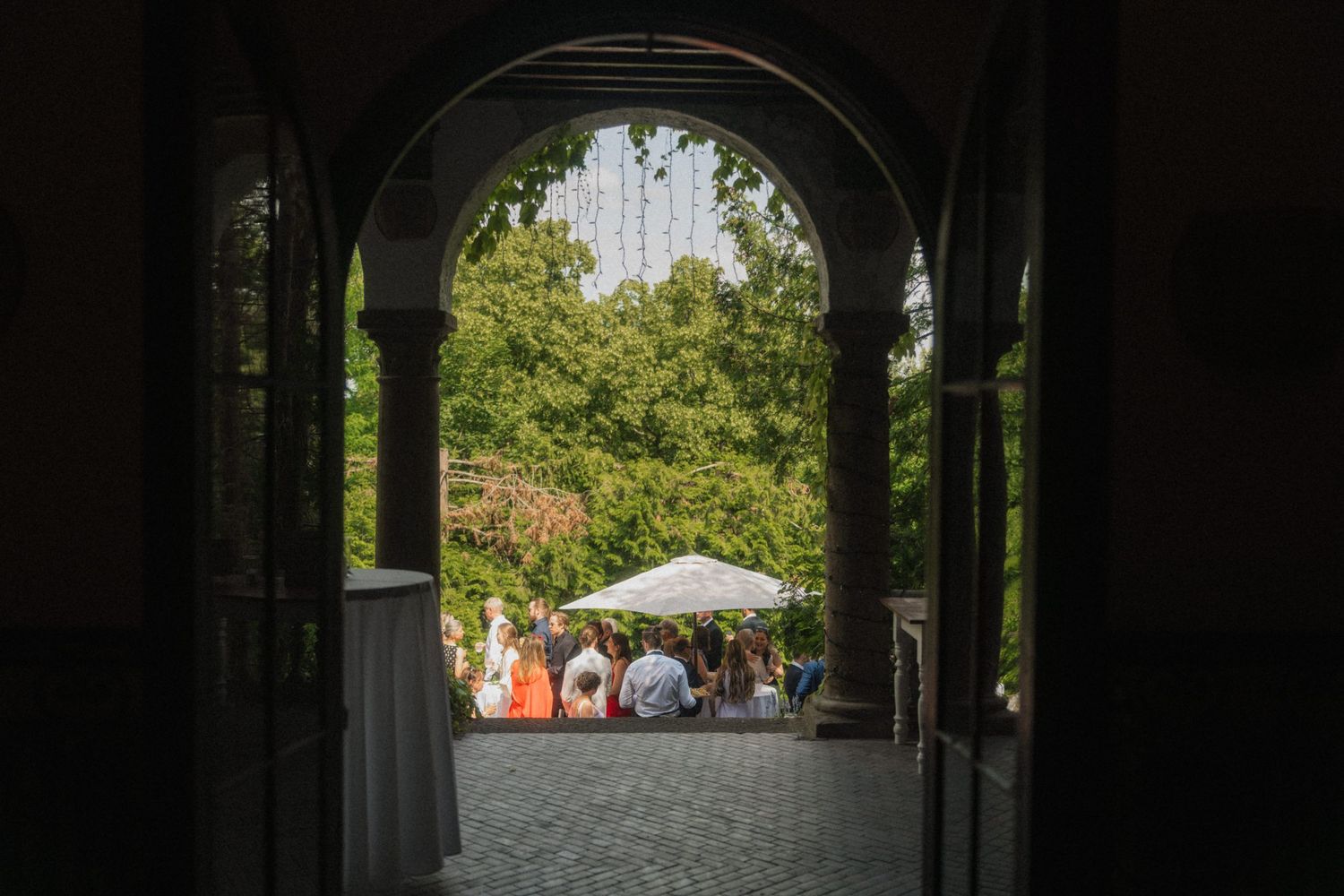 View through arched doorway of wedding guests gathering outside under trees on cobblestone courtyard.