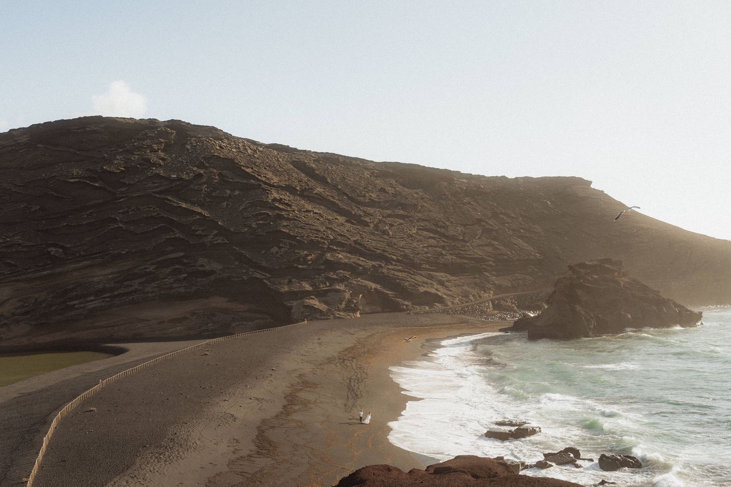 Scenic coastal landscape with black sand beach, rolling waves, and dramatic volcanic hillside at sunset.