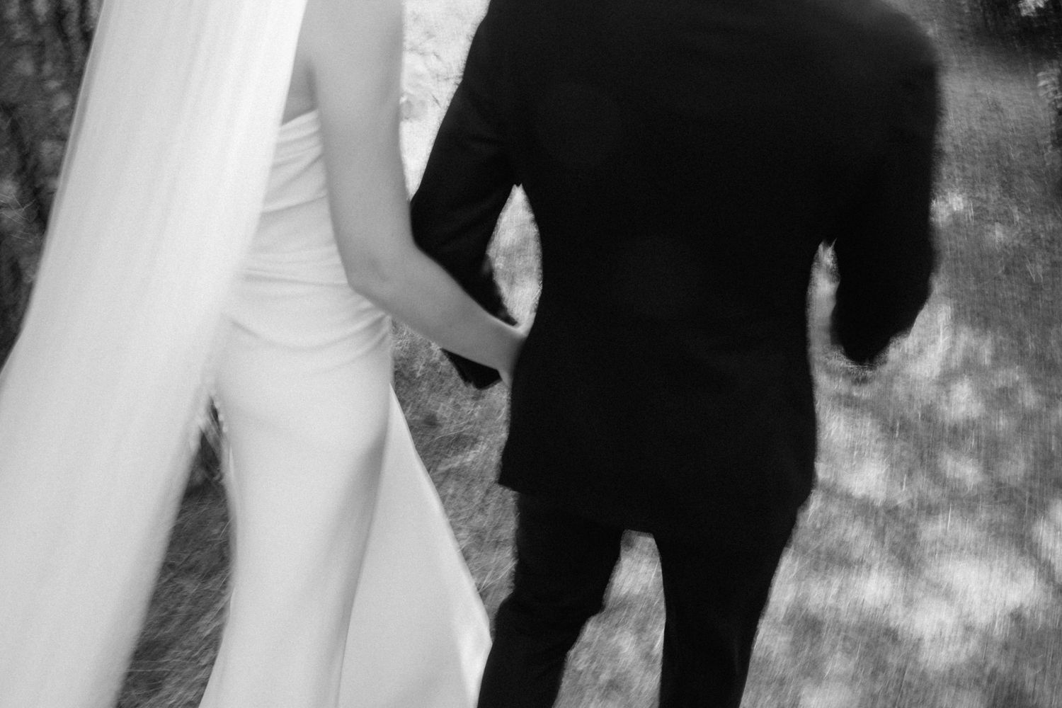 Black and white photo of a bride and groom holding hands, walking together outdoors on their wedding day.
