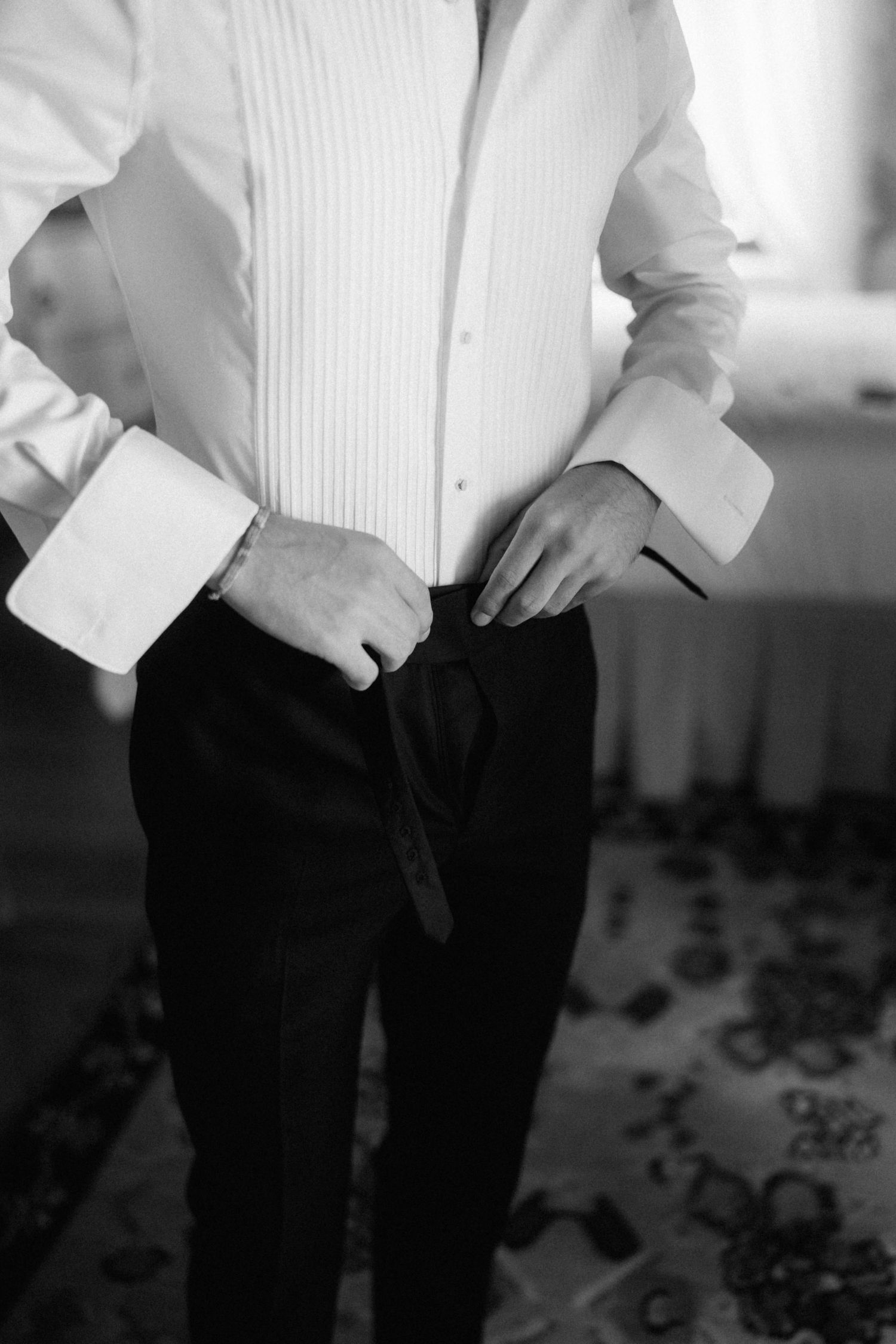 Groom getting dressed in white tuxedo shirt and black trousers on wedding day, black and white.