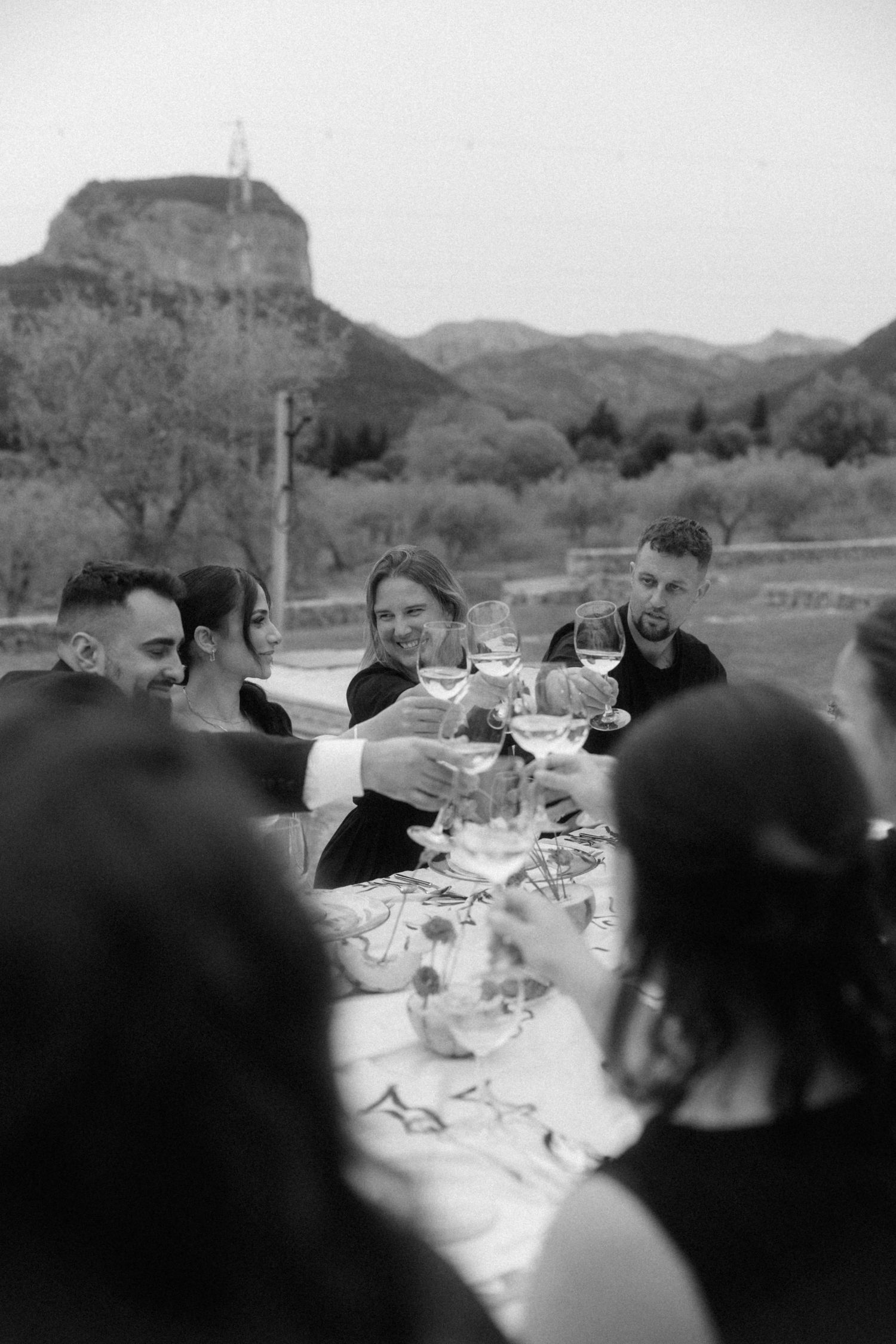 Wedding guests raise glasses in a toast at outdoor reception table with mountain views in black and white.