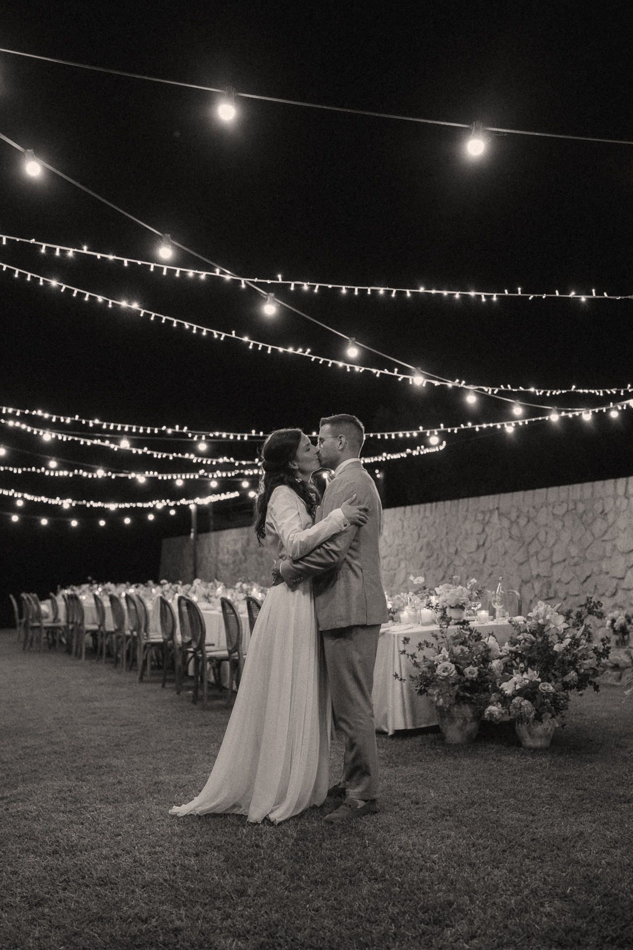Couple kissing under string lights at outdoor wedding reception at night, black and white photo.