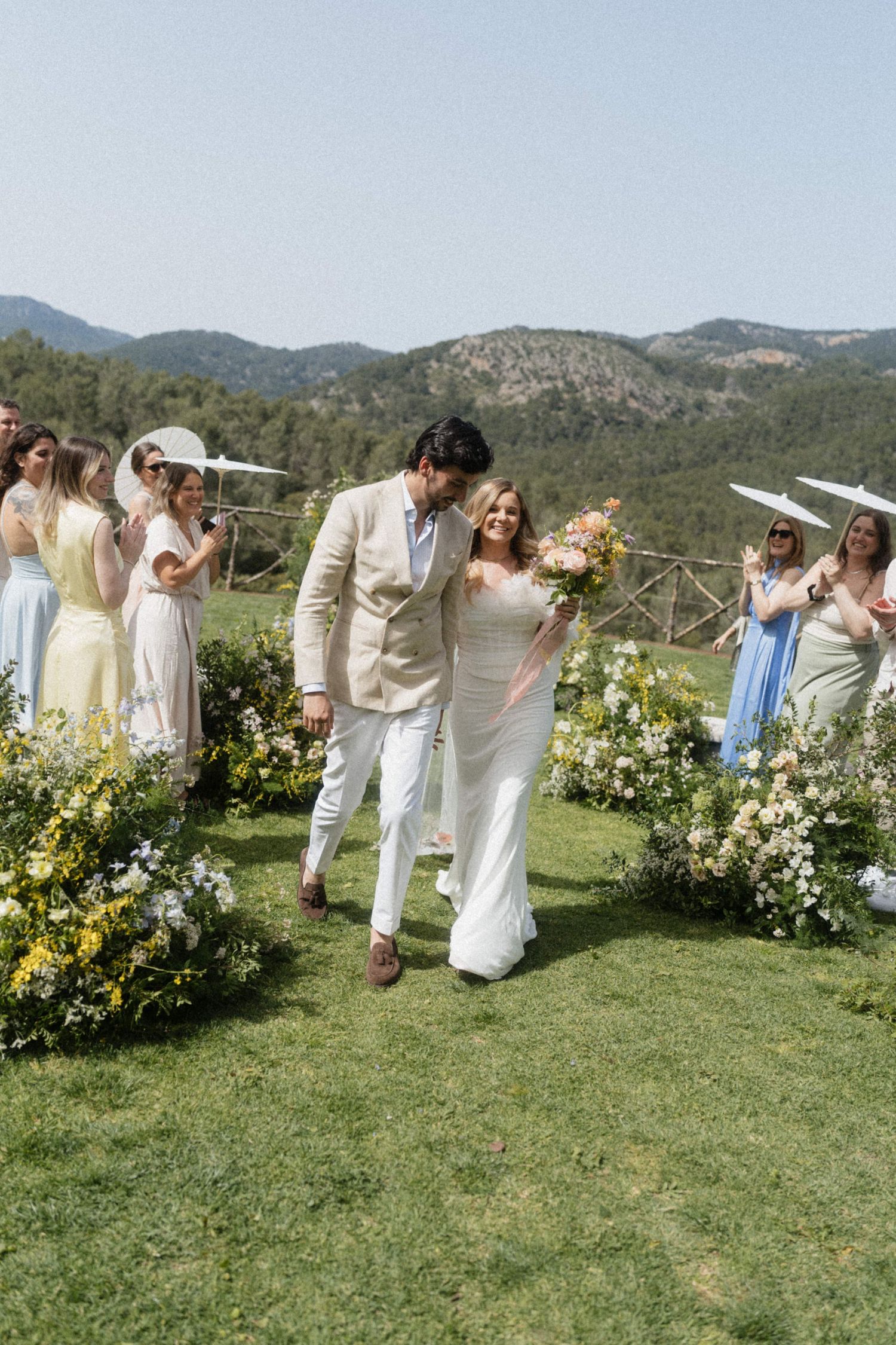 Bride and groom walk down flower-lined aisle at outdoor wedding ceremony surrounded by guests with parasols.
