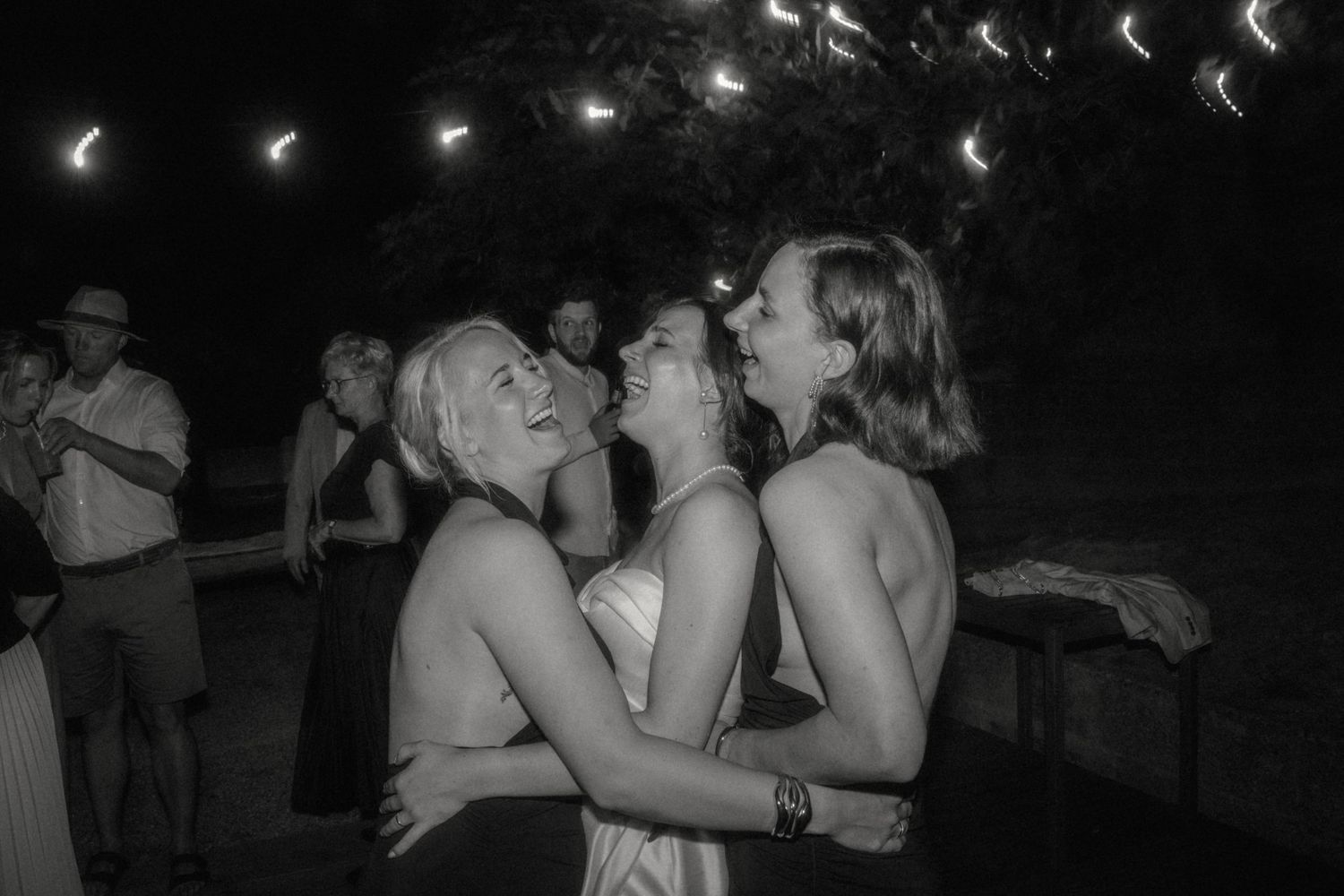 Black and white photo of bride and female guests laughing and dancing together at outdoor wedding reception.