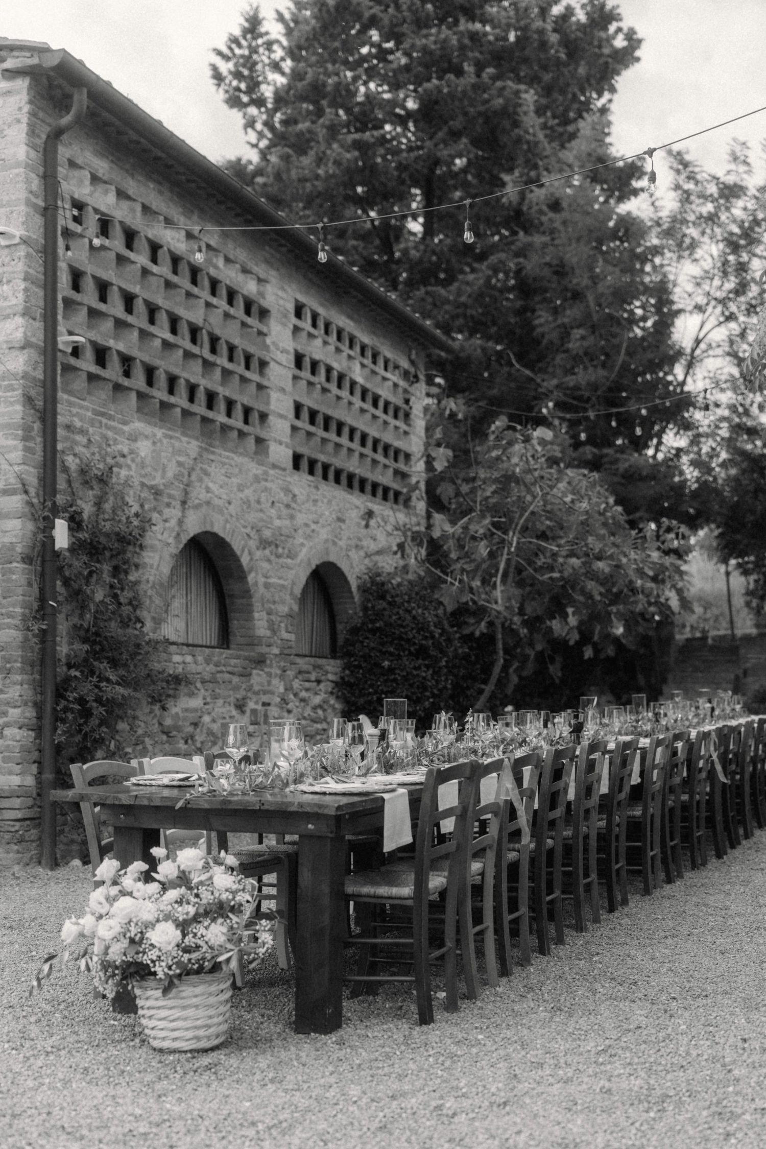 Black and white photo of long outdoor wedding banquet table beside rustic Italian stone building.